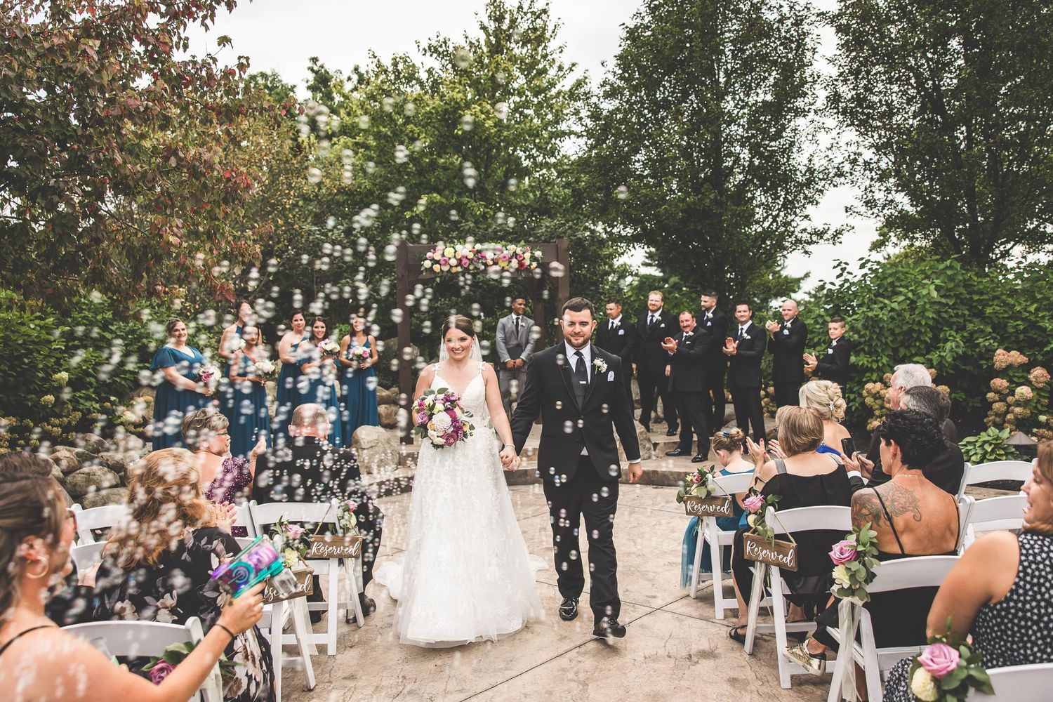 Wide view of wedding ceremony showing guests seated in rows with white chairs outdoors.