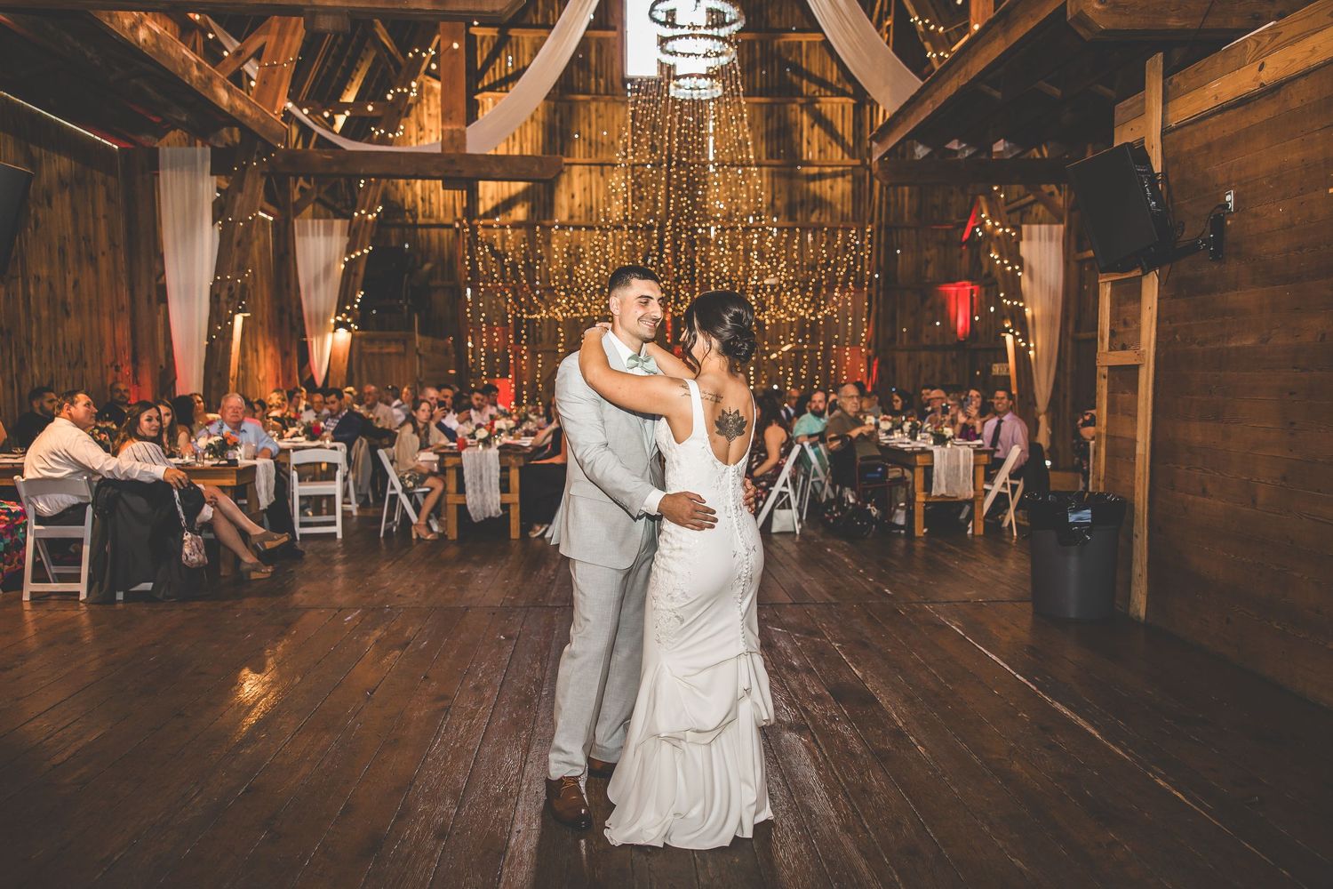 A couple shares their first dance in a rustic barn venue with twinkling lights and chandeliers overhead.
