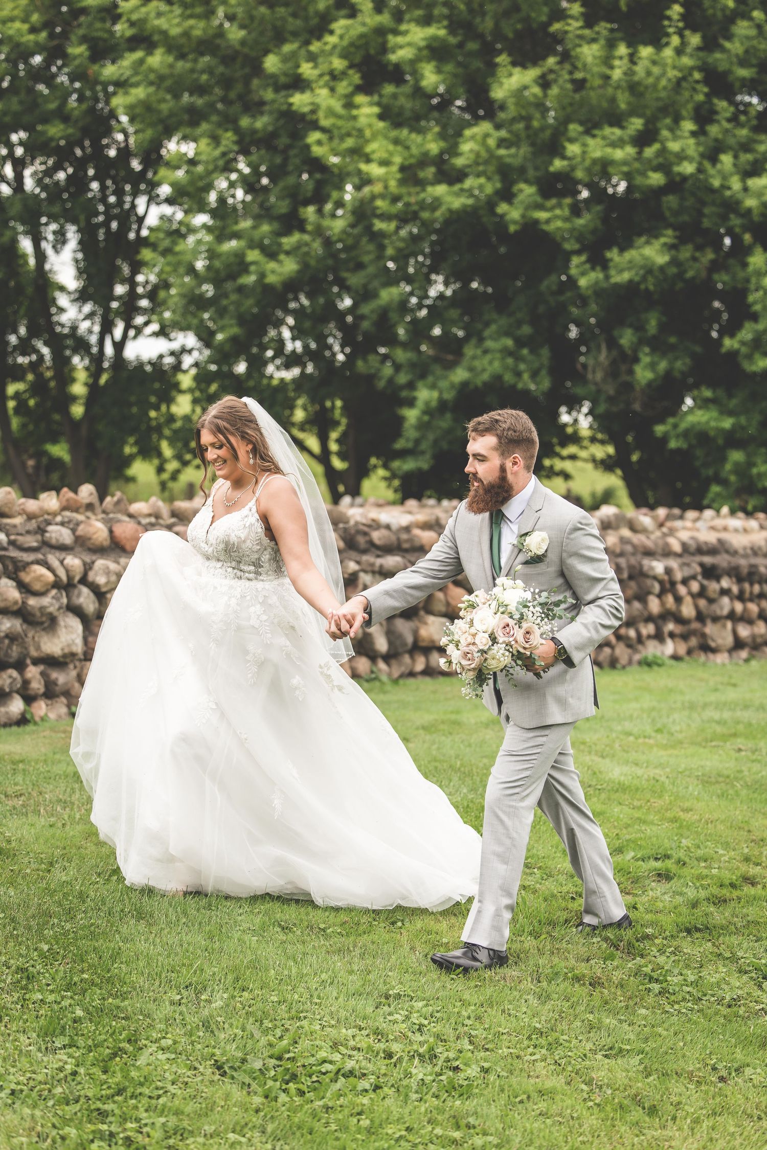 A playful wedding couple poses on grass near a stone wall as the bride's dress billows in the wind.