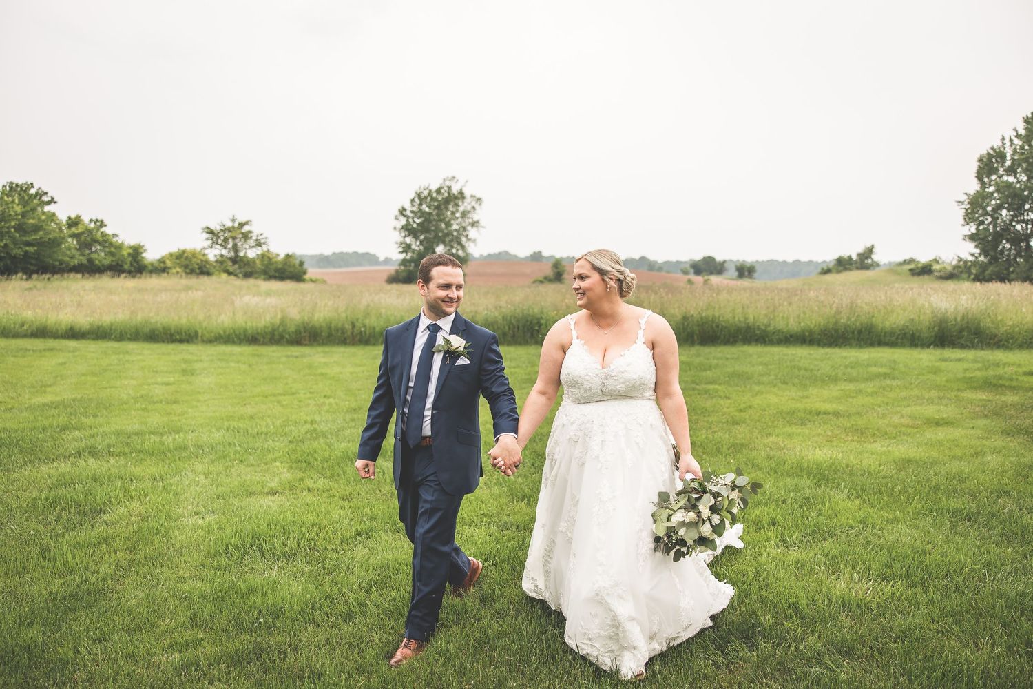 Newlyweds walk hand in hand through a scenic rural field on their wedding day.