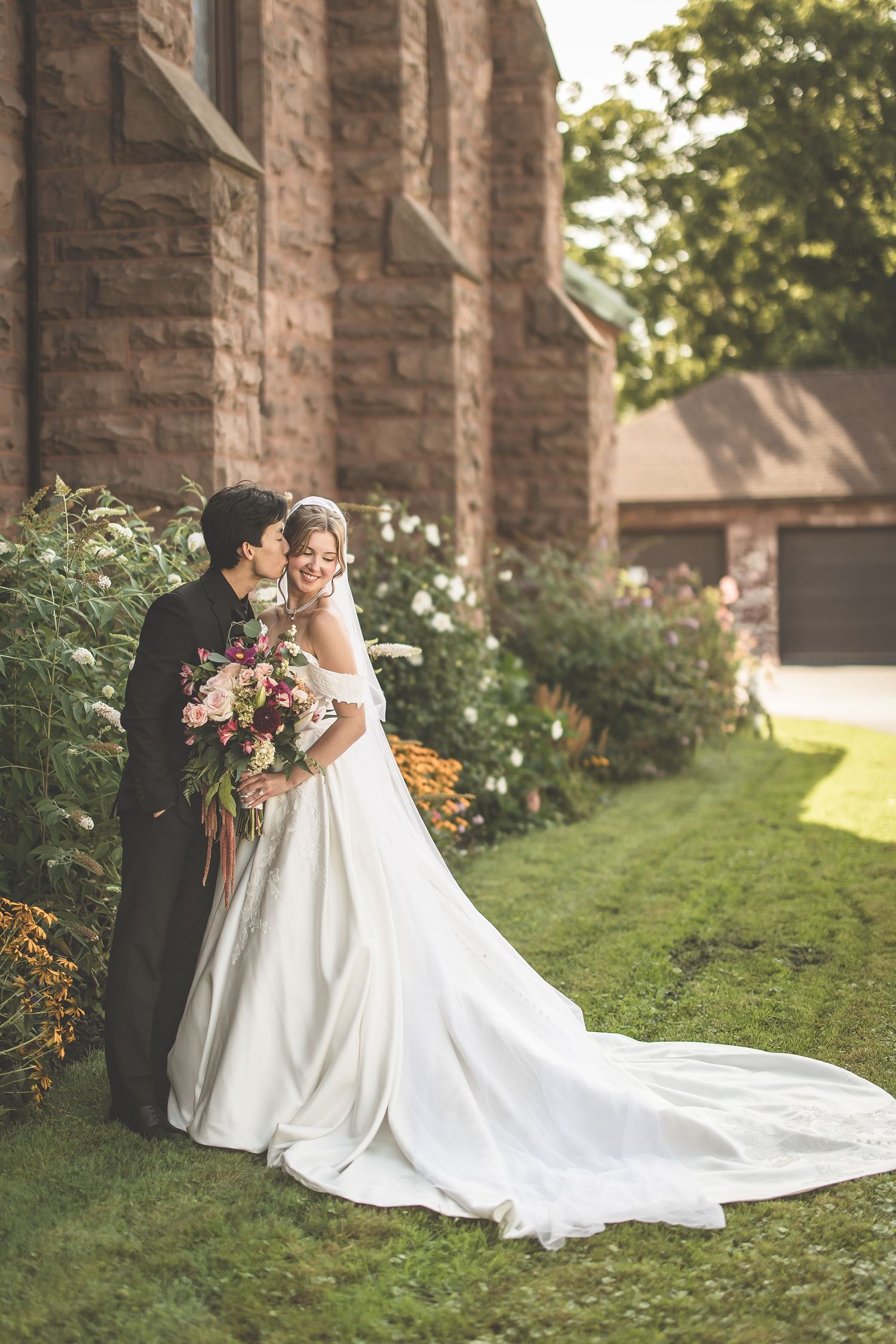 Bride and groom share a romantic moment outside a stone church, with bride holding a lush floral bouquet in a white gown.