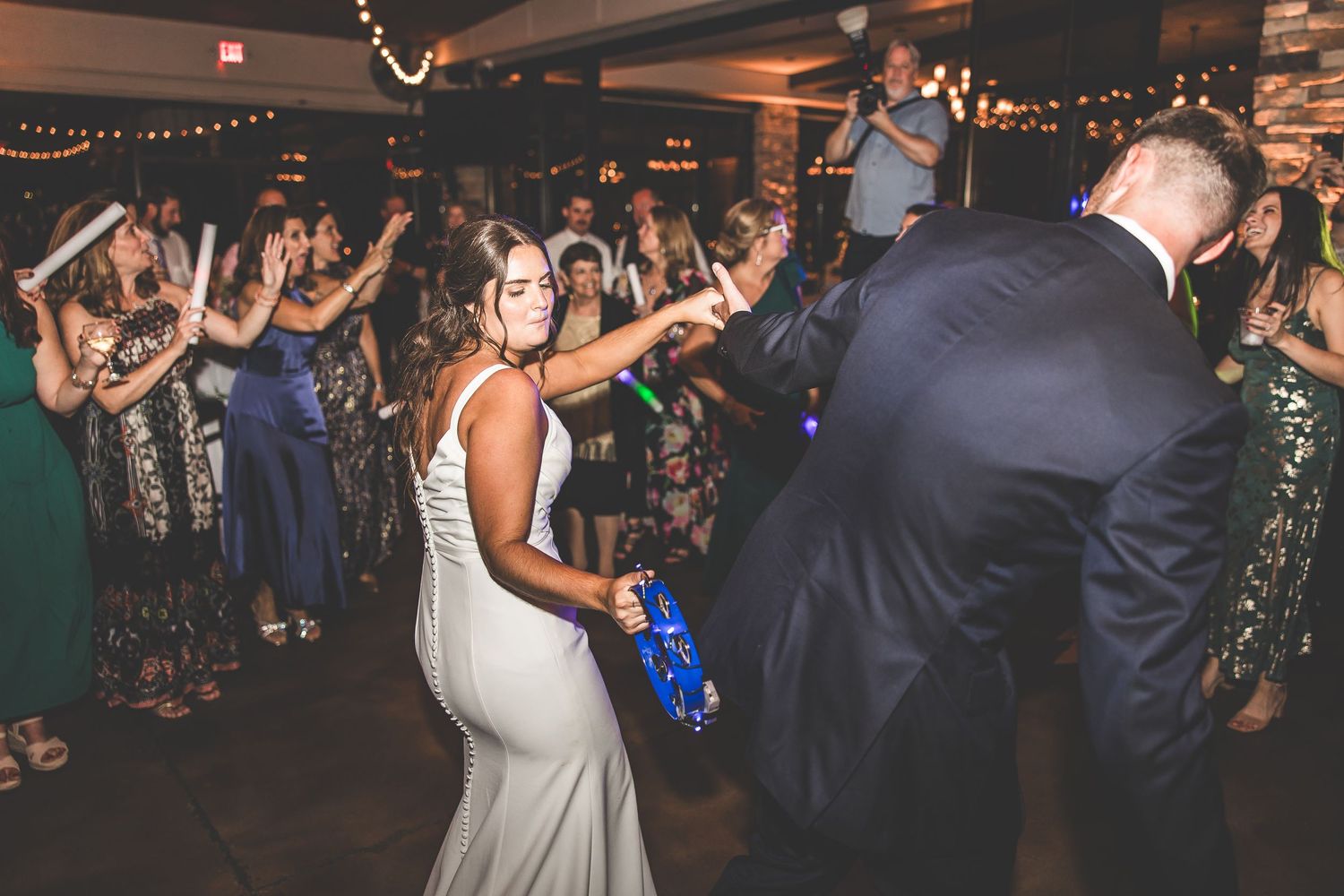 Wedding celebration with guests dancing under string lights in a rustic indoor venue.