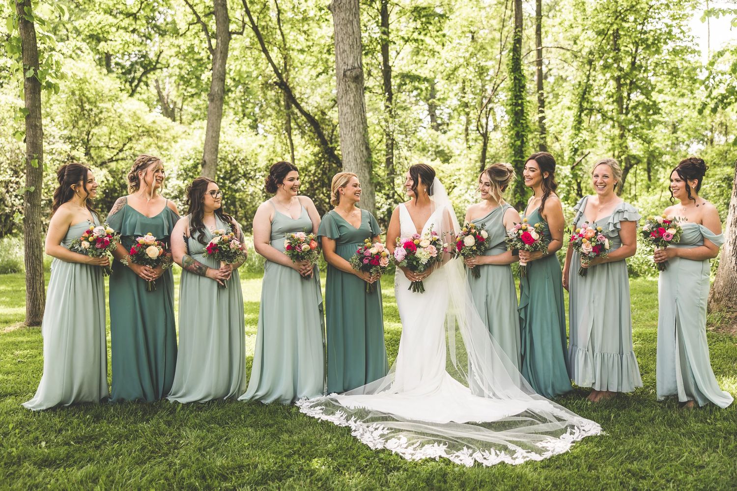 Bridesmaids in sage green dresses stand with the bride in a wooded setting.