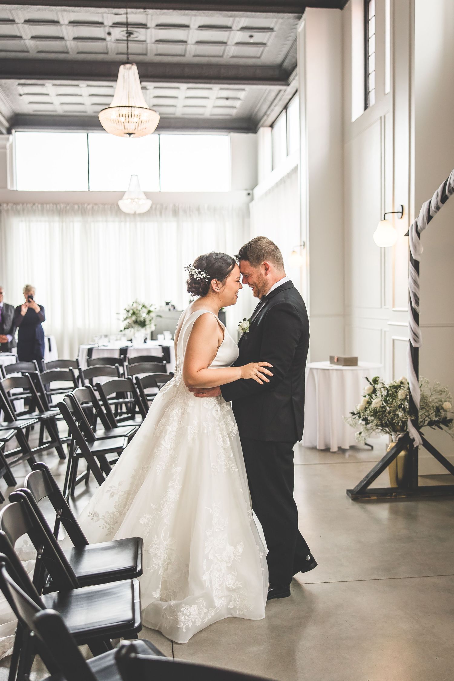 Wedding couple sharing first dance in elegant indoor venue with black chairs and white decor.