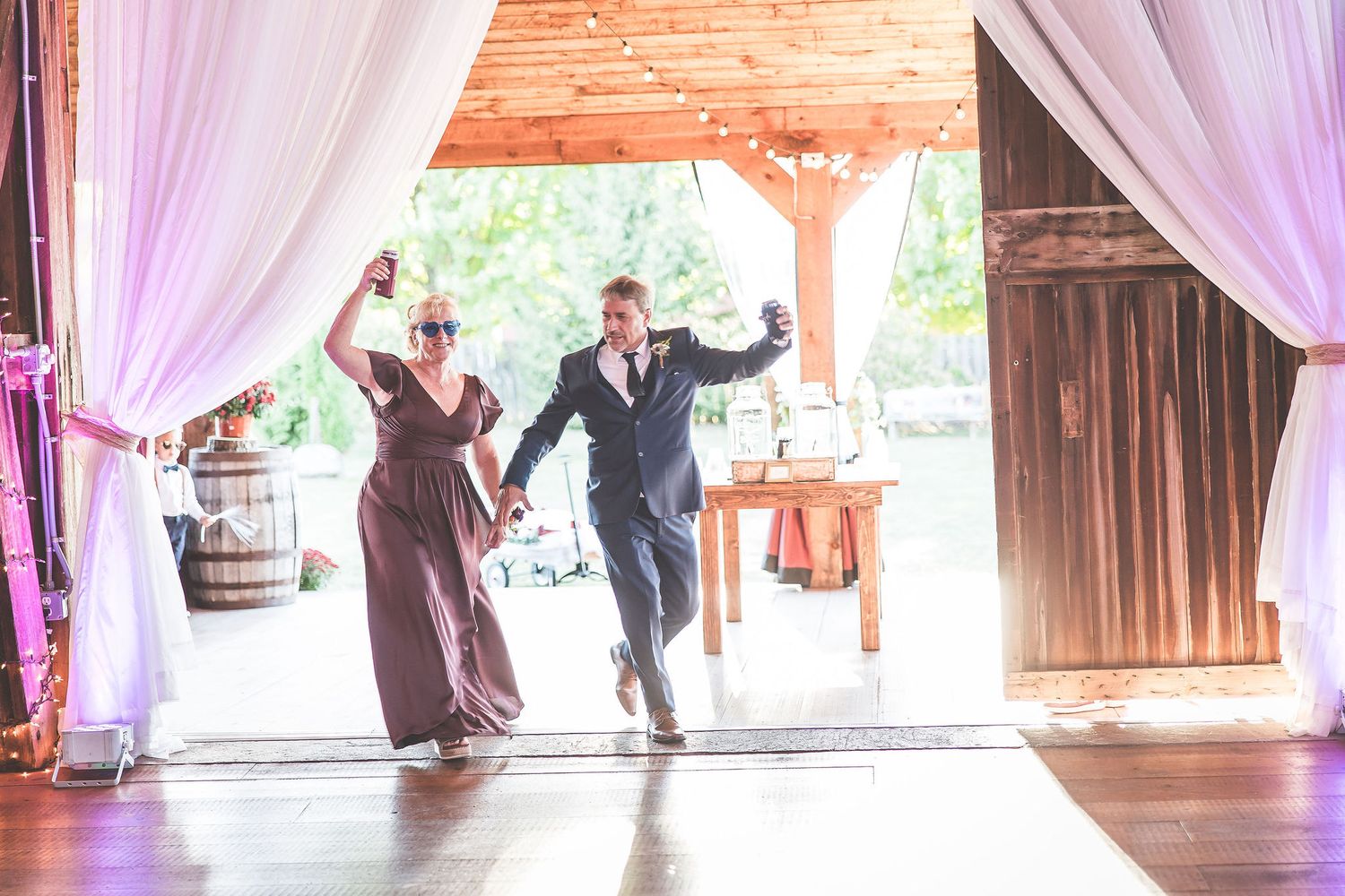 Wedding guests dancing together on wooden floor of barn venue decorated with flowing white fabric.