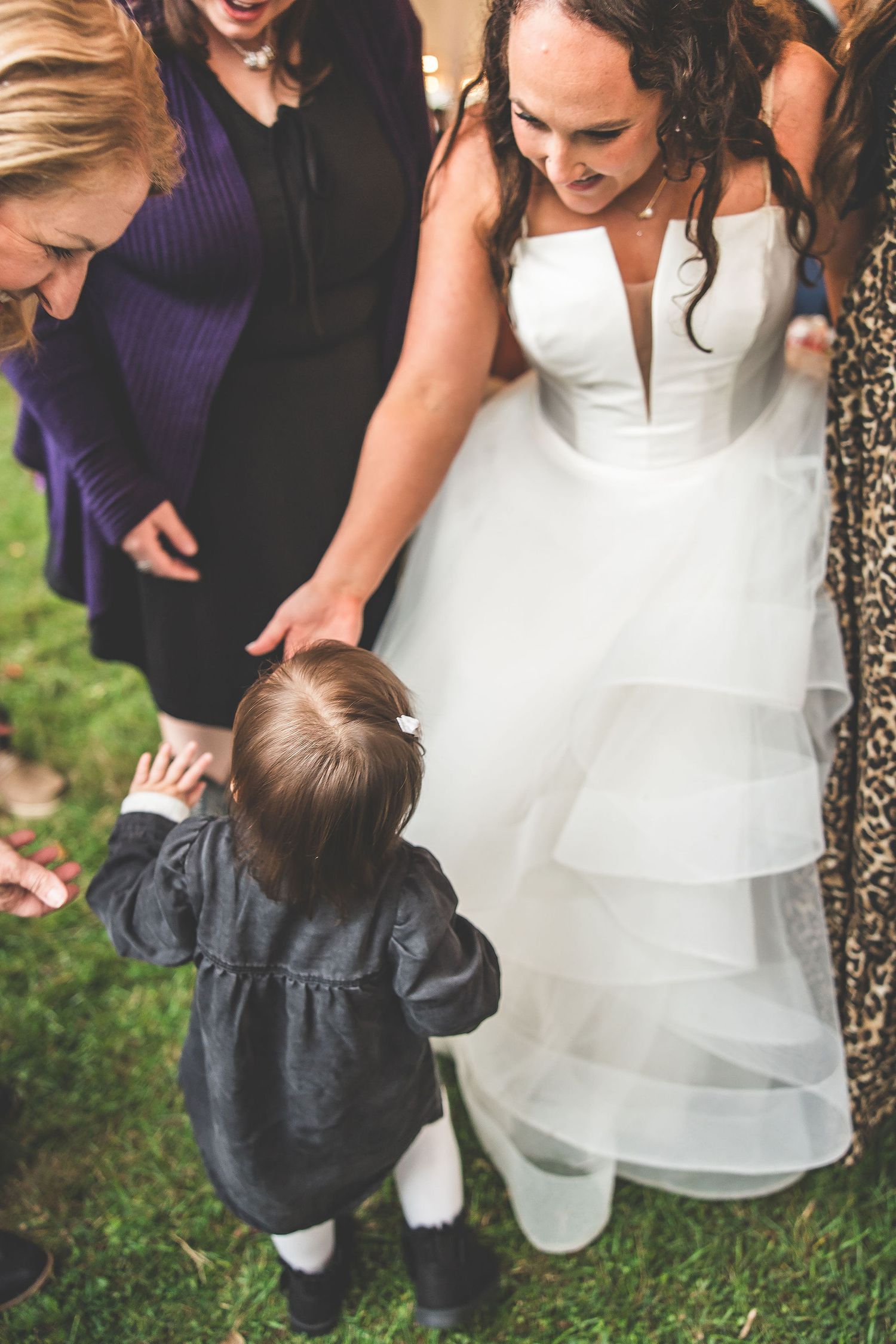 Small child in navy outfit crouches near white wedding dress during outdoor celebration.