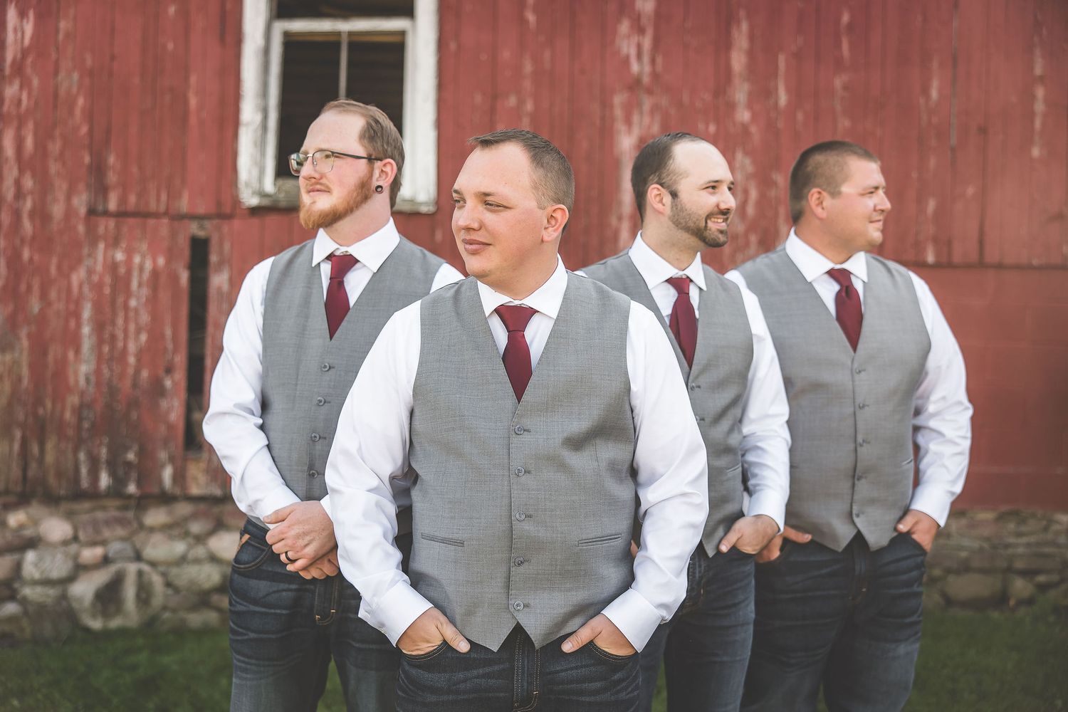 Groomsmen in gray vests and white shirts pose casually by rustic barn.