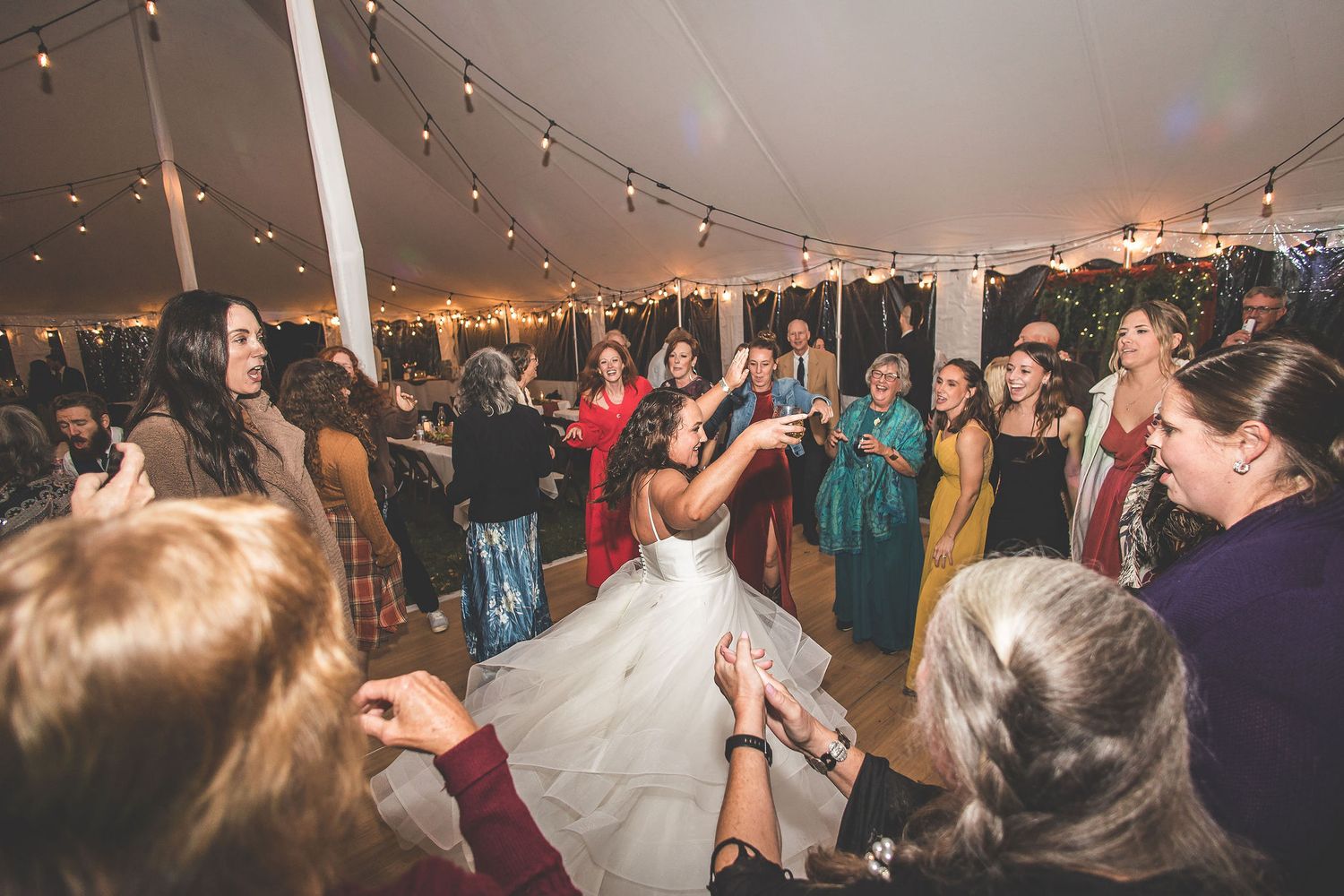 Wedding reception tent filled with dancing guests under glowing string lights.