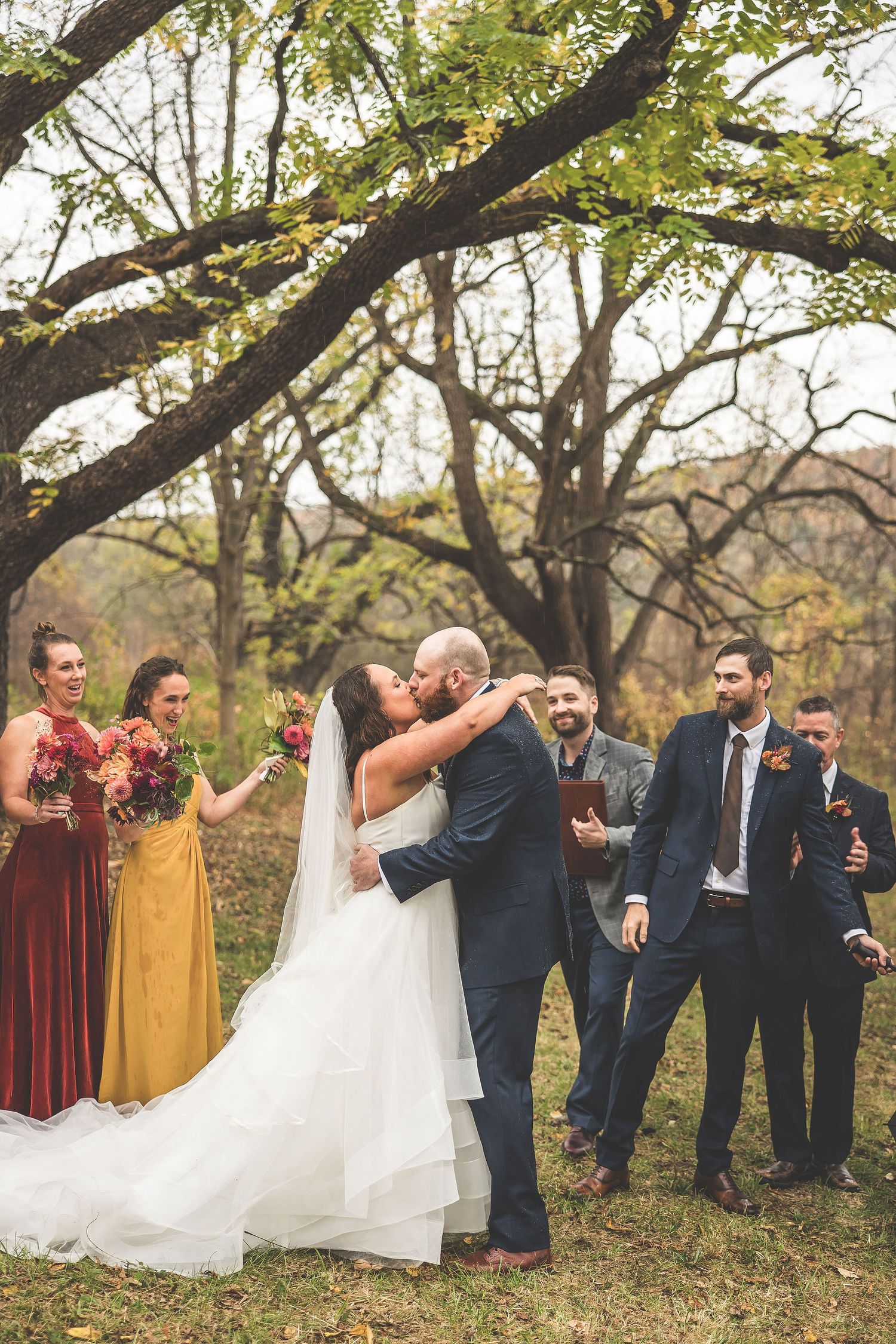 Newlyweds share first kiss while wedding party celebrates in autumn outdoor setting.