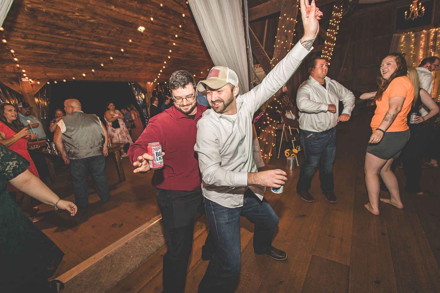 Party guests dance enthusiastically on a wooden floor under string lights at a reception celebration.