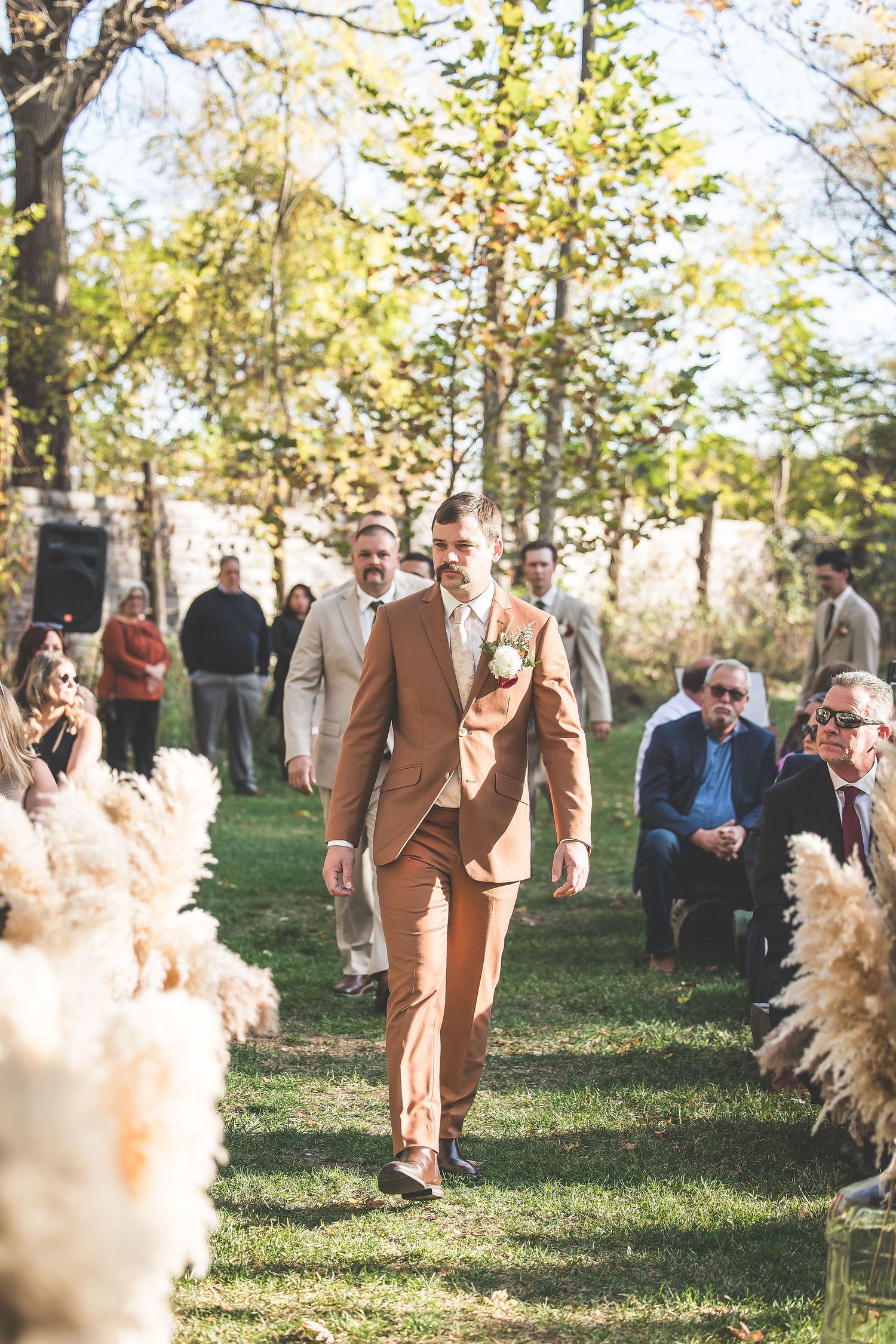 Wedding party member in brown suit walks down aisle lined with pampas grass and autumn trees.