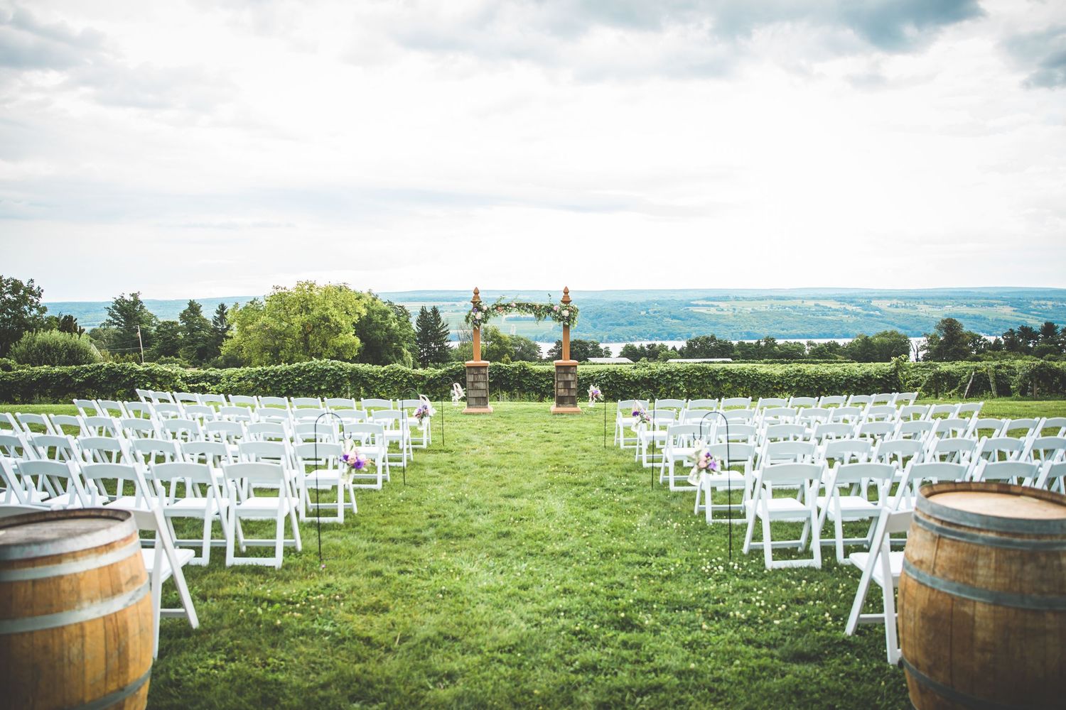 Outdoor wedding ceremony setup with white chairs and wine barrels on a rolling green hillside.