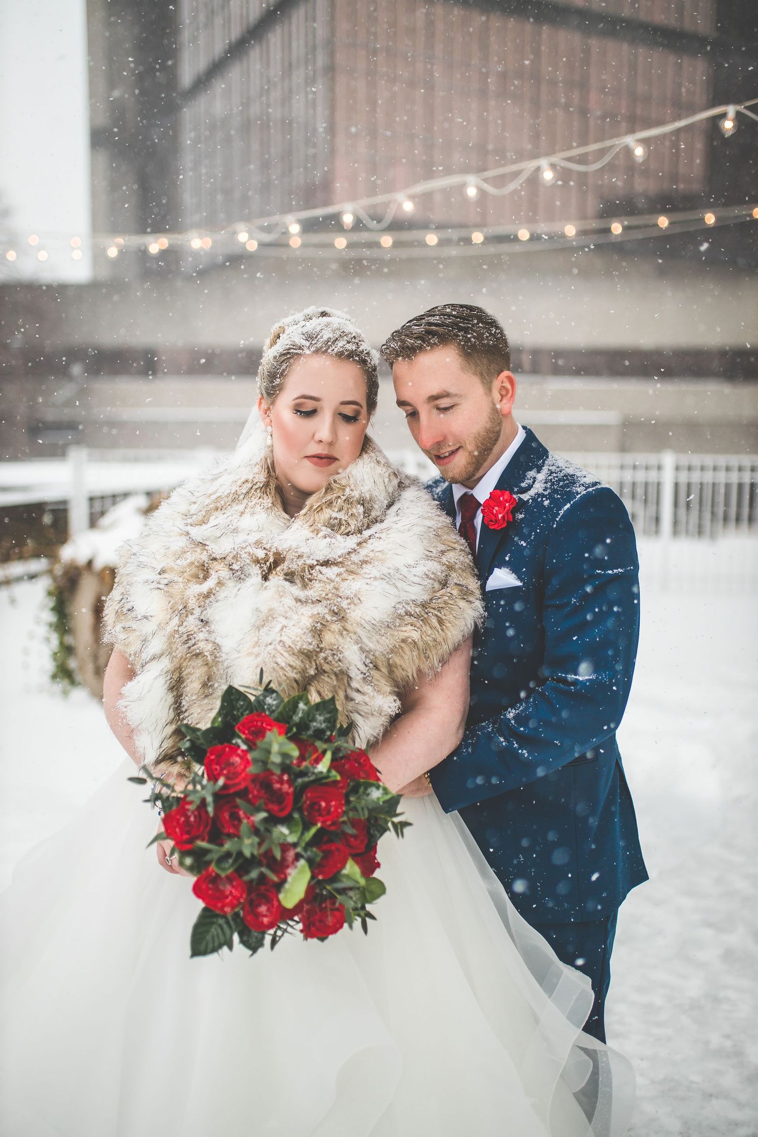 Winter wedding couple embraces in snowfall with bride wearing fur stole and holding red bouquet.