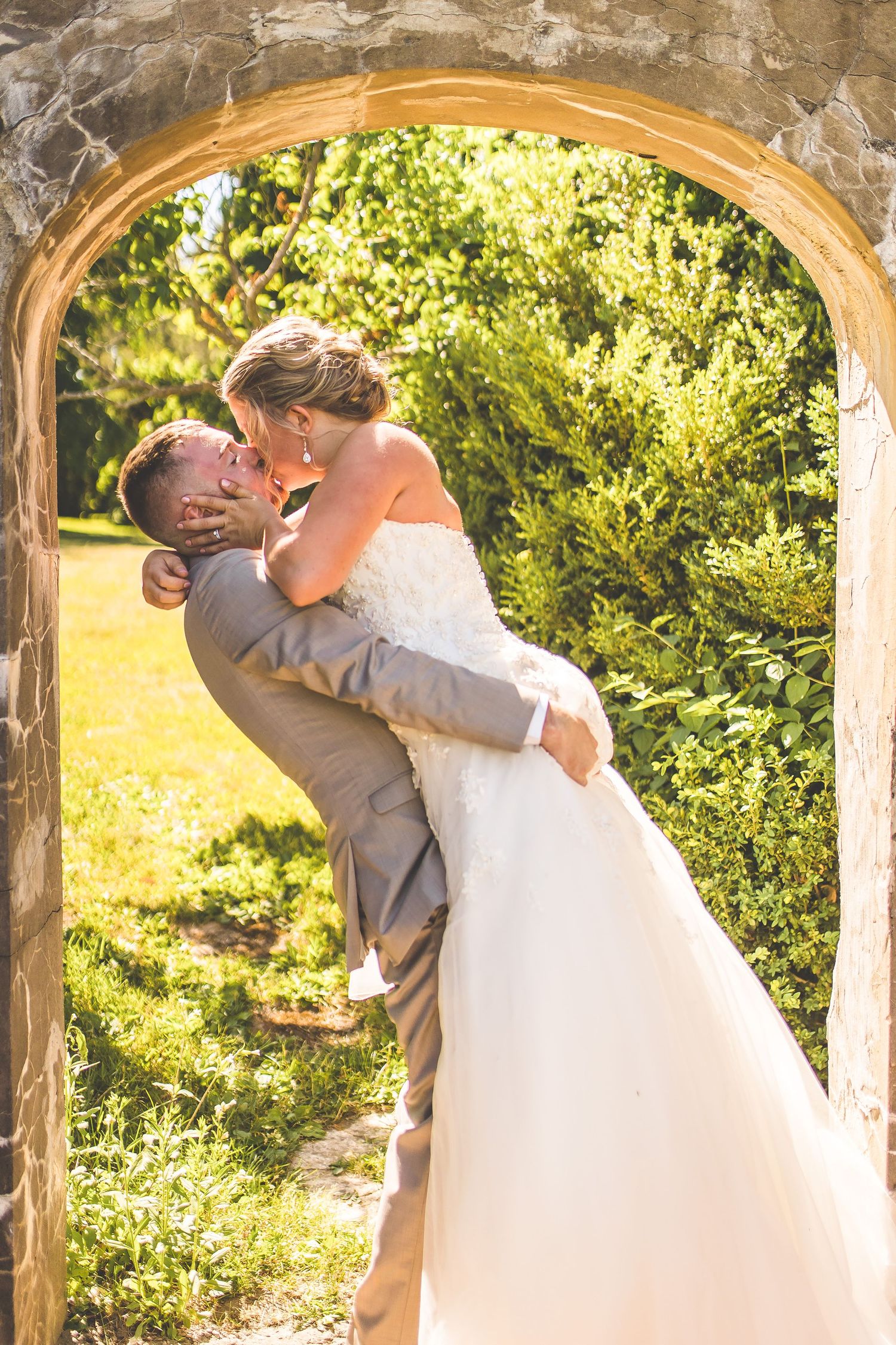 Wedding couple embraces under a rustic stone archway in golden afternoon light.