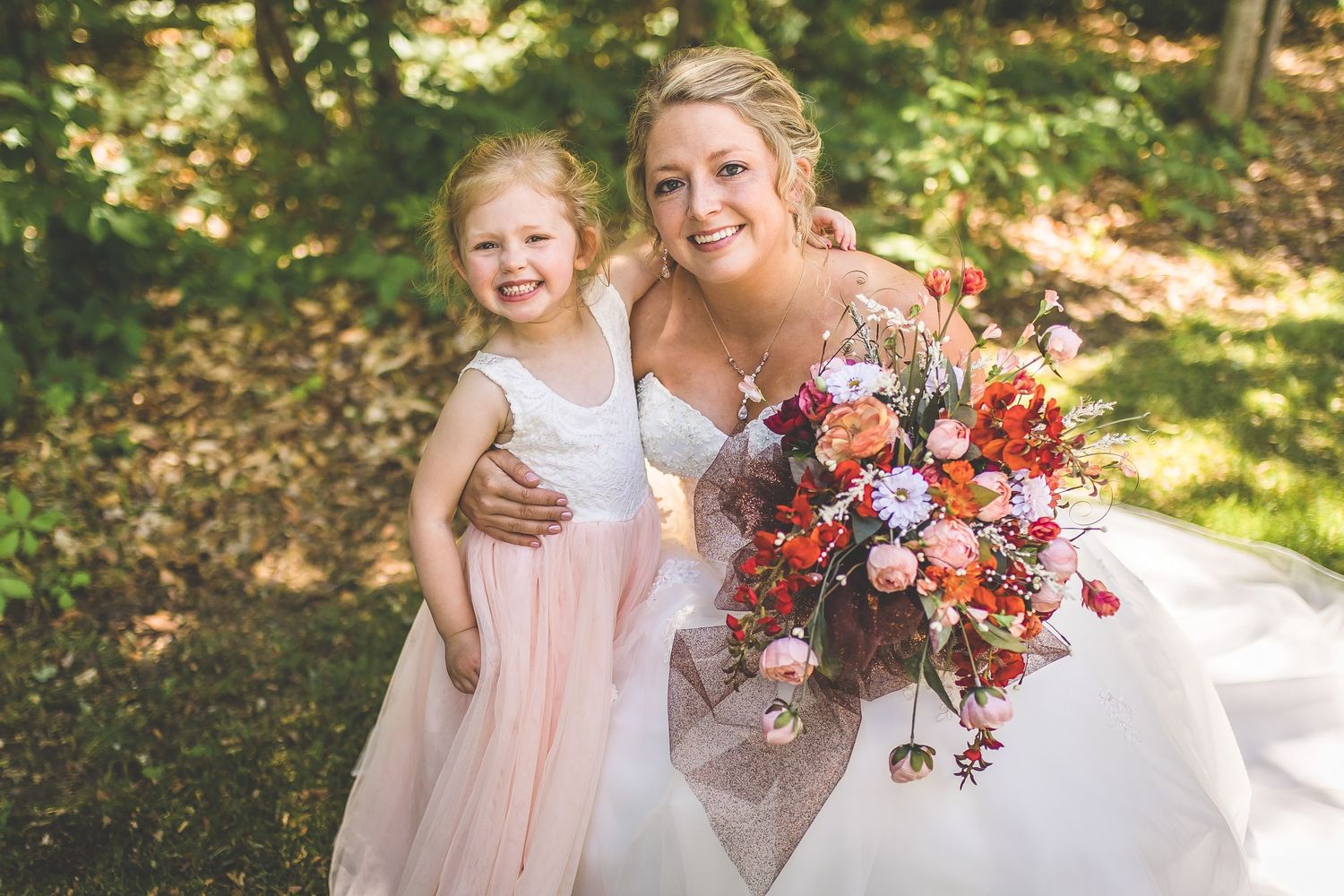 Bridesmaids in blush pink dresses gather outdoors holding vibrant fall-colored bouquets at a woodland wedding.