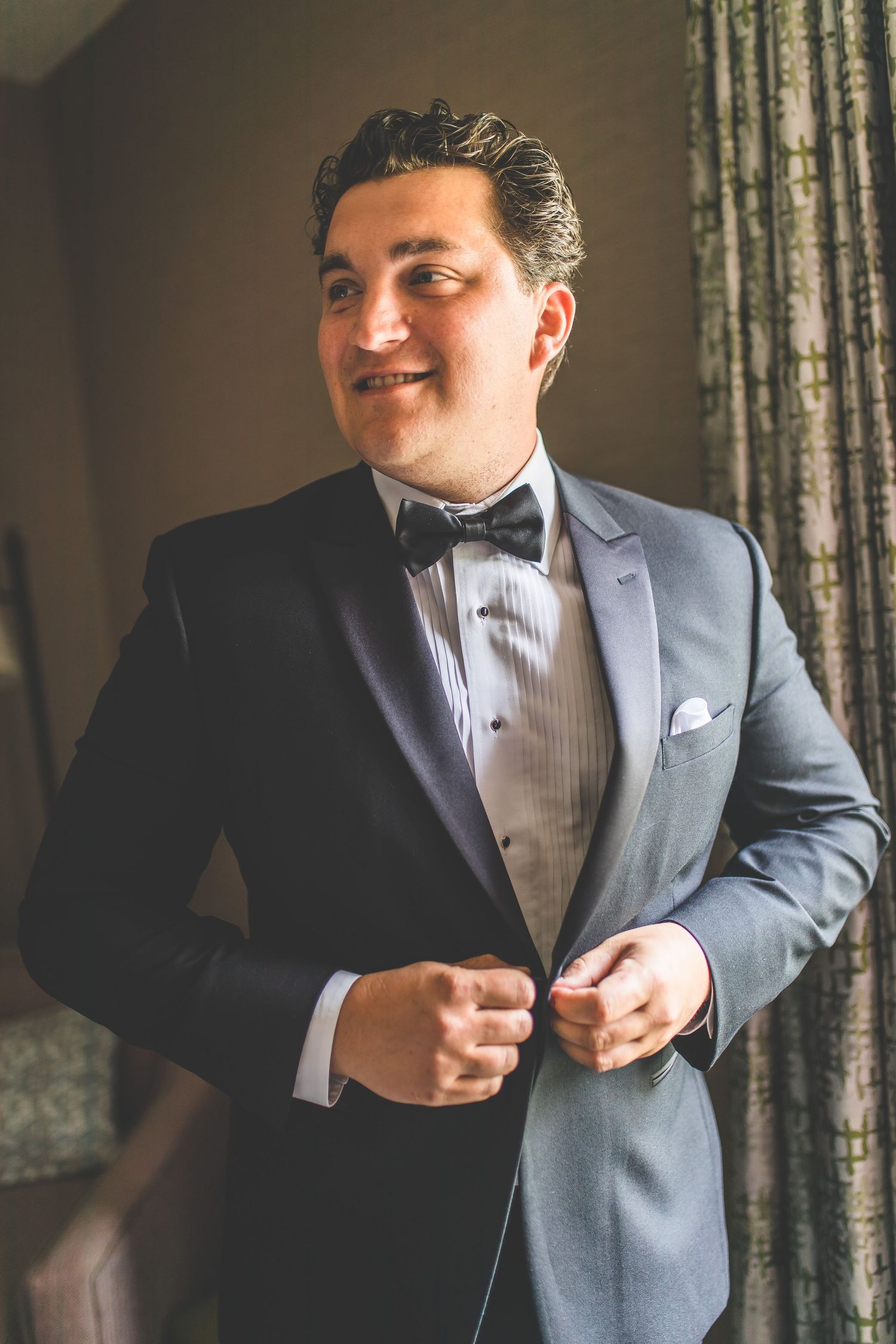 Groom adjusting his bowtie while getting ready in grey tuxedo.