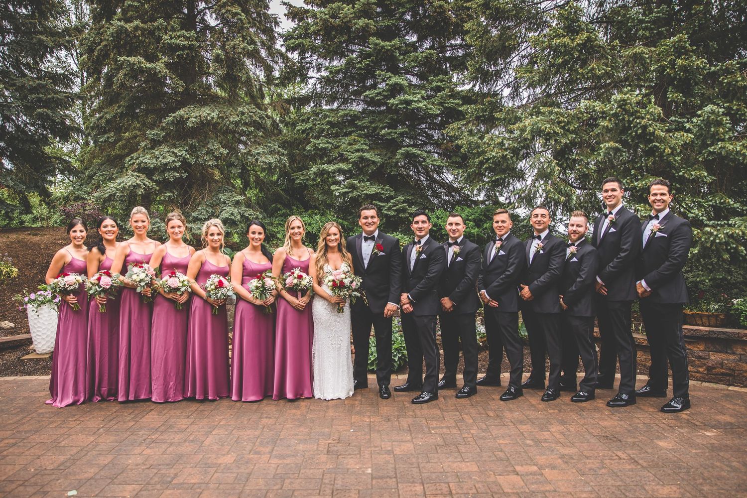 Full wedding party portrait with bridesmaids in rose dresses and groomsmen in black suits on brick patio.