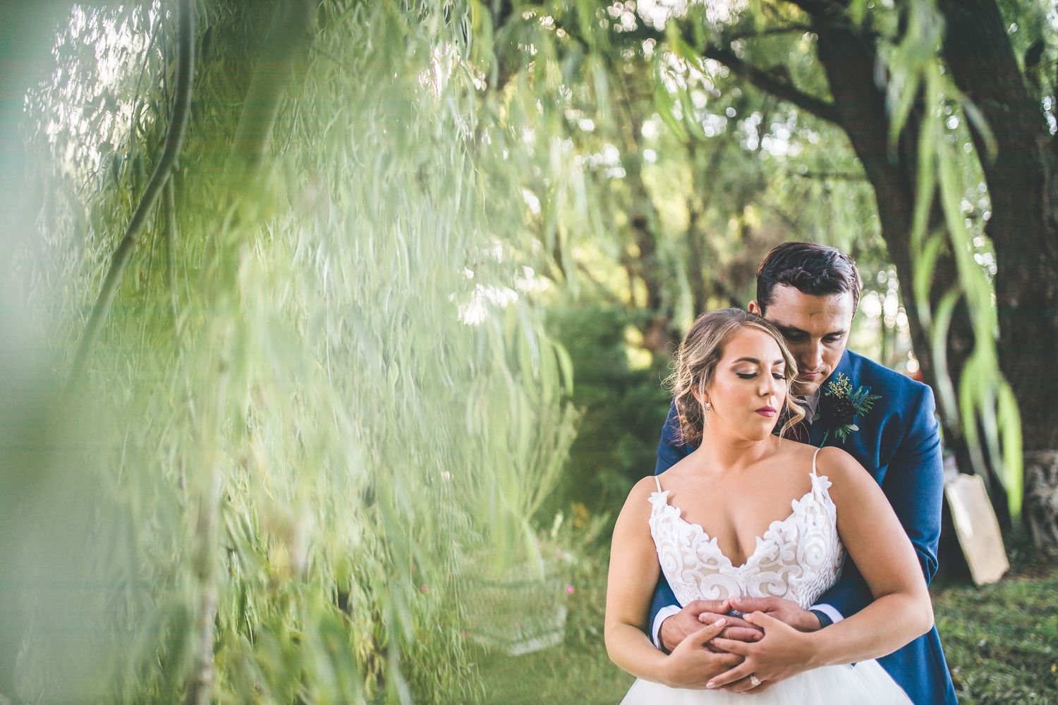Romantic portrait of couple embracing under weeping willow trees with soft green lighting.