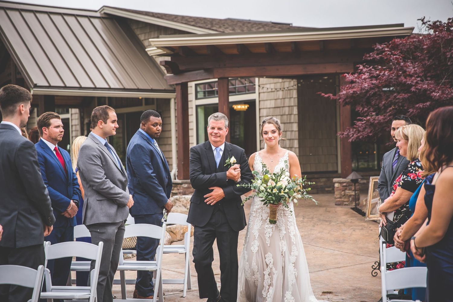 Outdoor wedding ceremony setup with white chairs and floral arrangements under a covered pavilion.