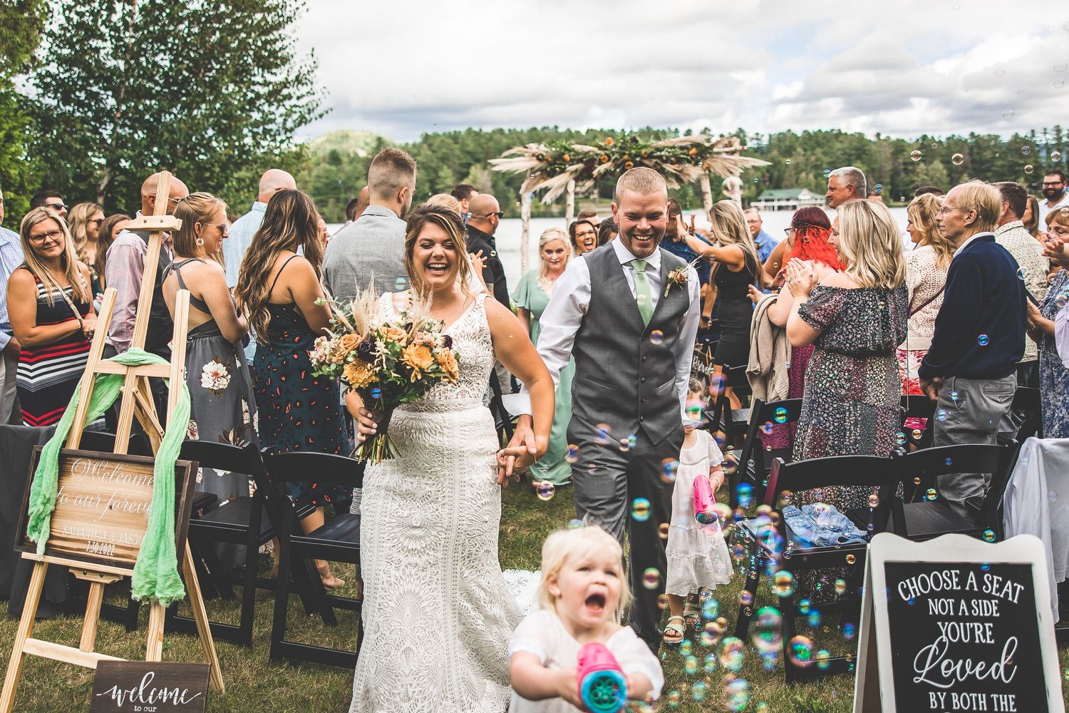 Joyful wedding celebration with guests and bubbles during outdoor lakeside ceremony.