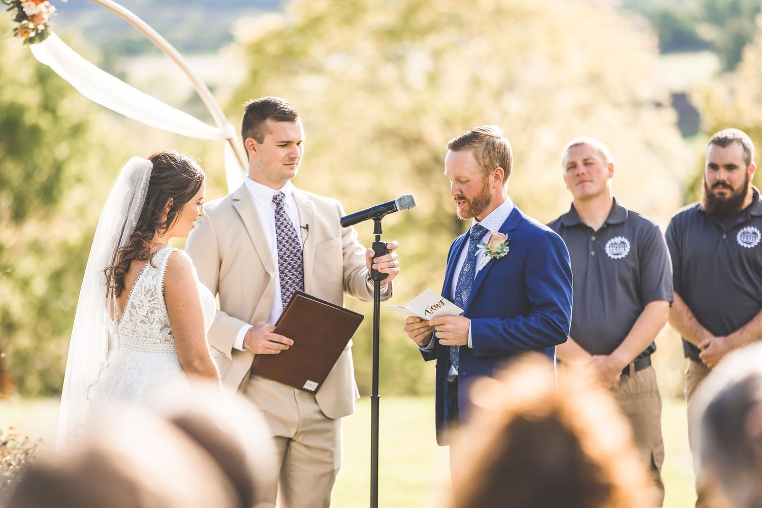 Outdoor wedding ceremony takes place under golden sunlight with guests seated on white chairs.