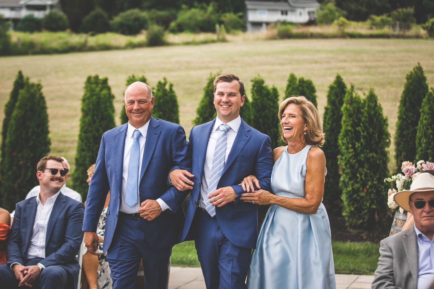 Wedding guests mingle on manicured lawn with cypress trees.