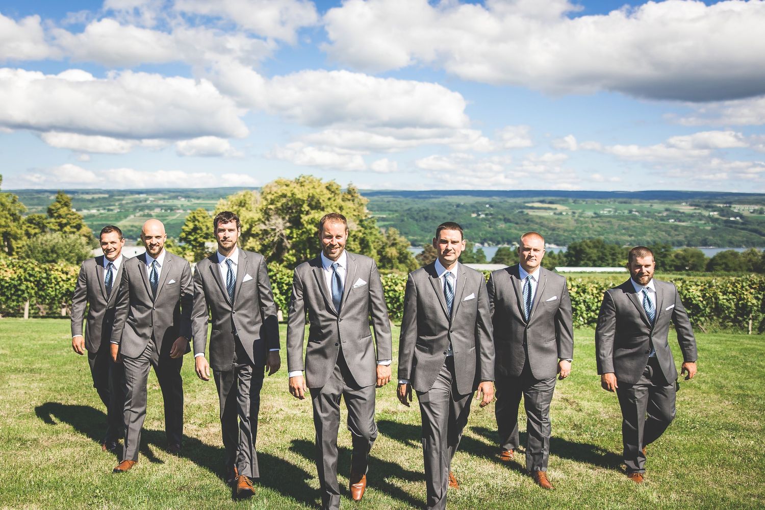 Groomsmen in gray suits walking together across a scenic hilltop with cloudy sky backdrop.