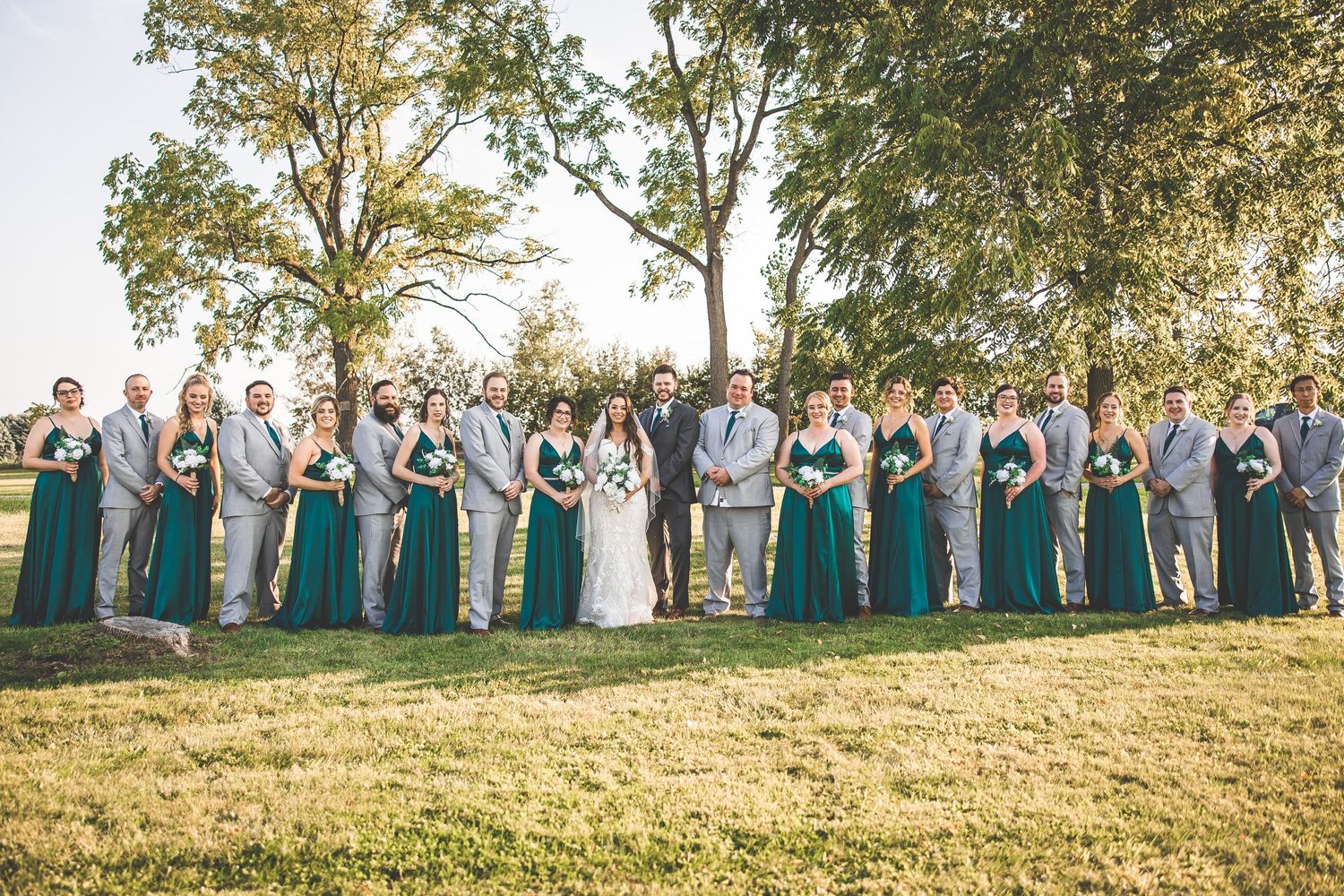 Wedding party in teal dresses stands in a line on a grassy hill during sunset.