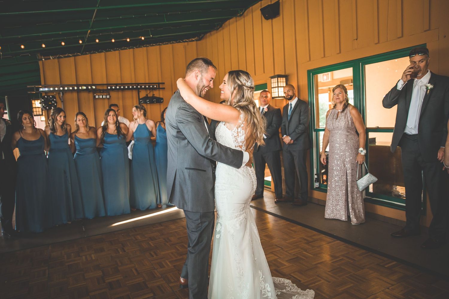 Wedding guests observe as newlyweds dance in an orange-walled venue with string lighting.