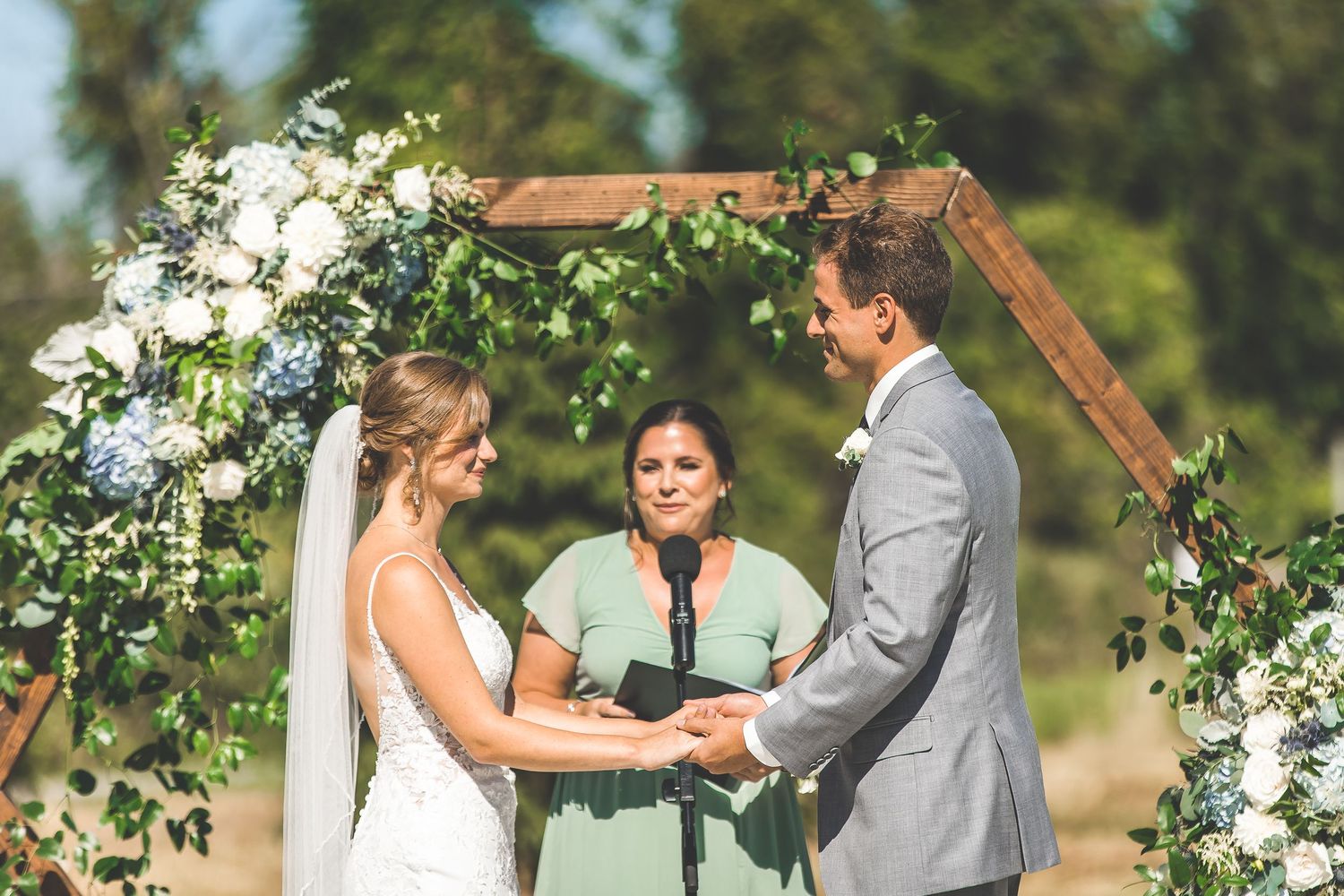 Wedding ceremony under wooden arch decorated with white flowers and greenery.