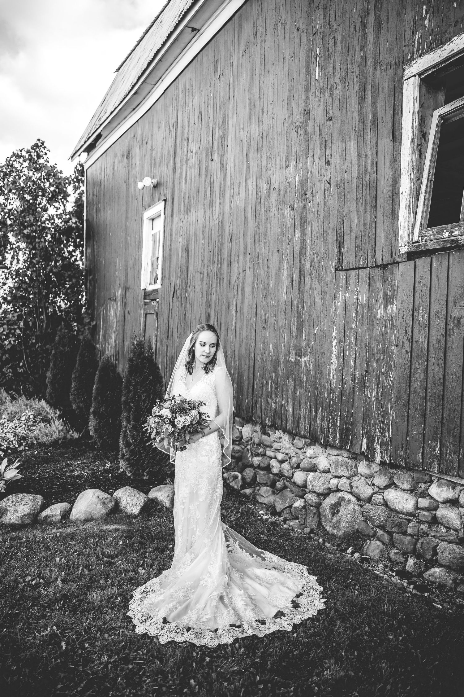 Black and white portraits of bride in lace gown against rustic barn wall and stone foundation.