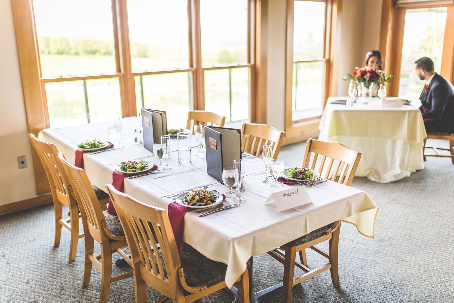 Light-filled reception venue with wooden chairs and white tablecloths set for wedding celebration.