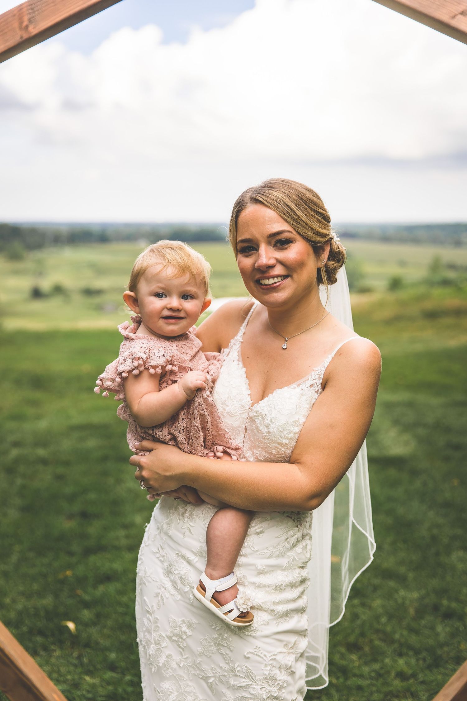 Bride in lace wedding dress holds small child in pink dress under wooden arch.