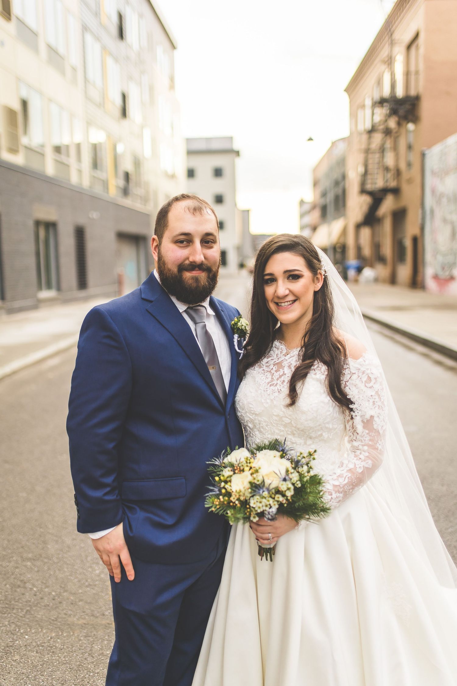 Newlywed couple poses on city street with bride in lace gown and groom in navy suit.