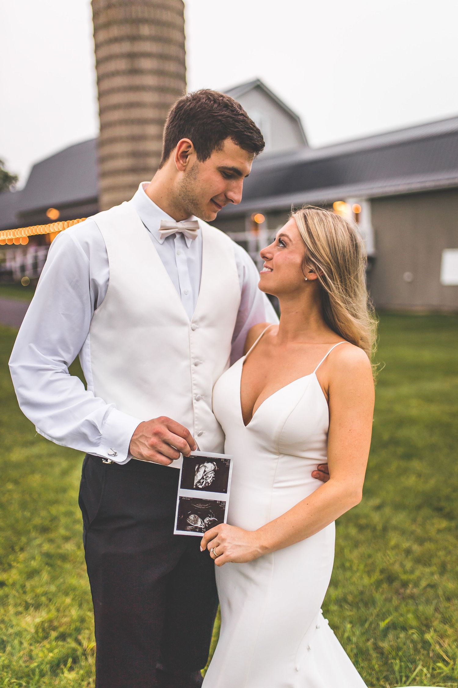 A couple shares an intimate moment outdoors while holding ultrasound photos during a pregnancy announcement photoshoot.