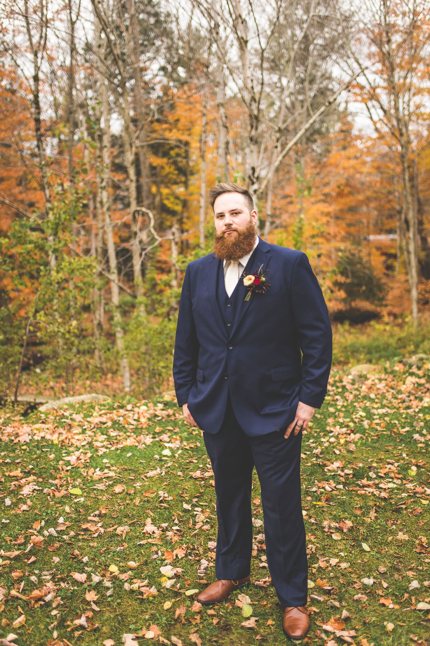 Groomsmen in navy suits pose casually on leaf-covered ground with fall foliage backdrop.