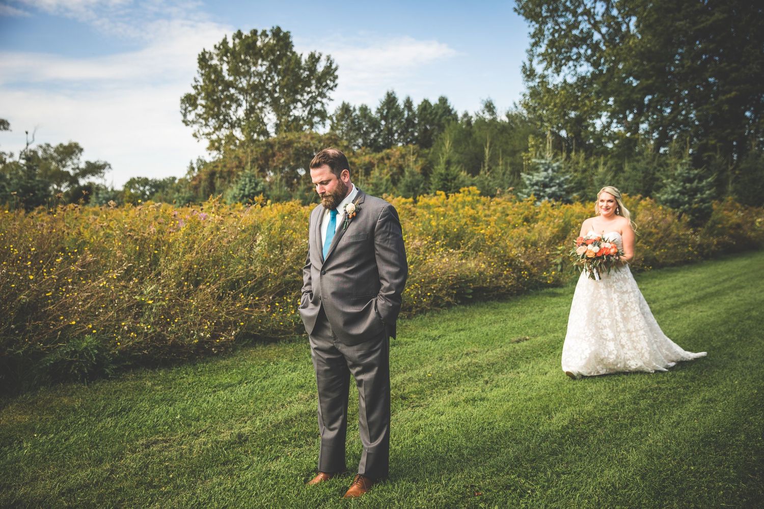 Wedding couple poses in a scenic outdoor field with yellow wildflowers and trees in the background.