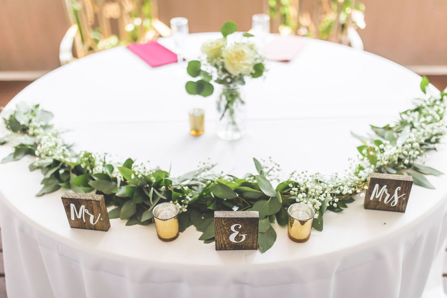 Elegant wedding table setting with white linens, greenery garland, and rustic wooden signs.