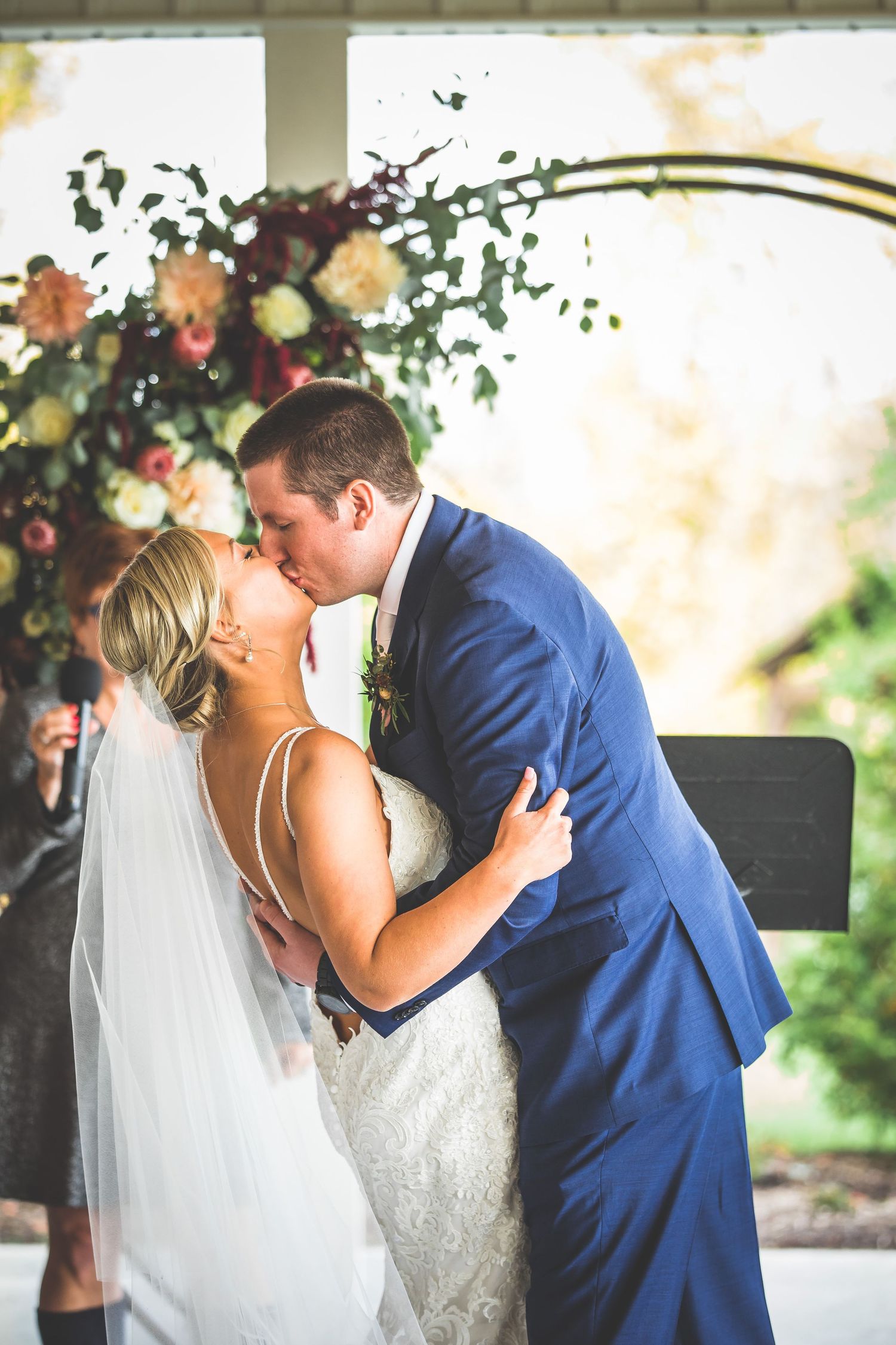 Romantic moment under floral arch decorated with peach and burgundy flowers at outdoor ceremony.