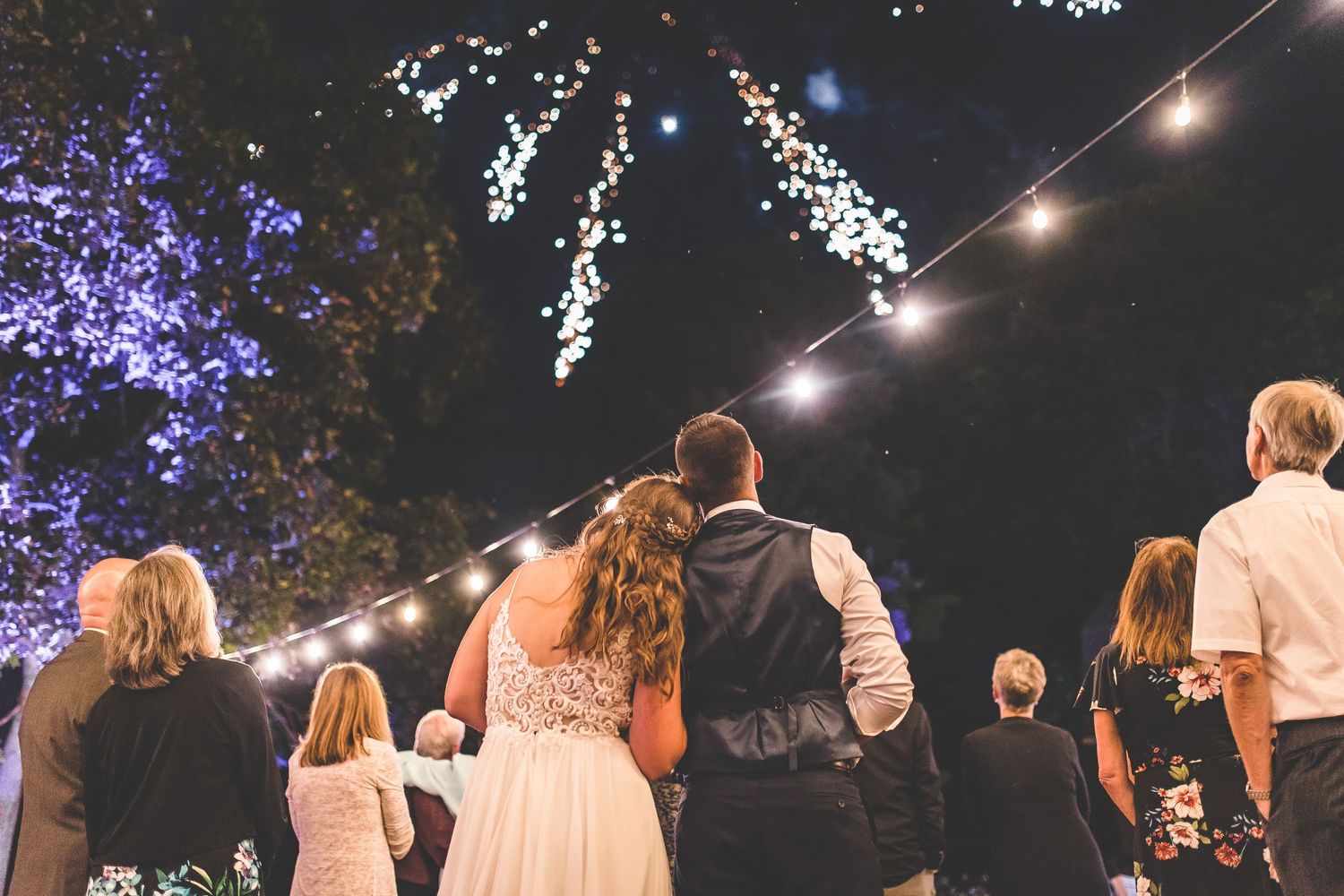 Wedding guests stand together watching fireworks display in the night sky.