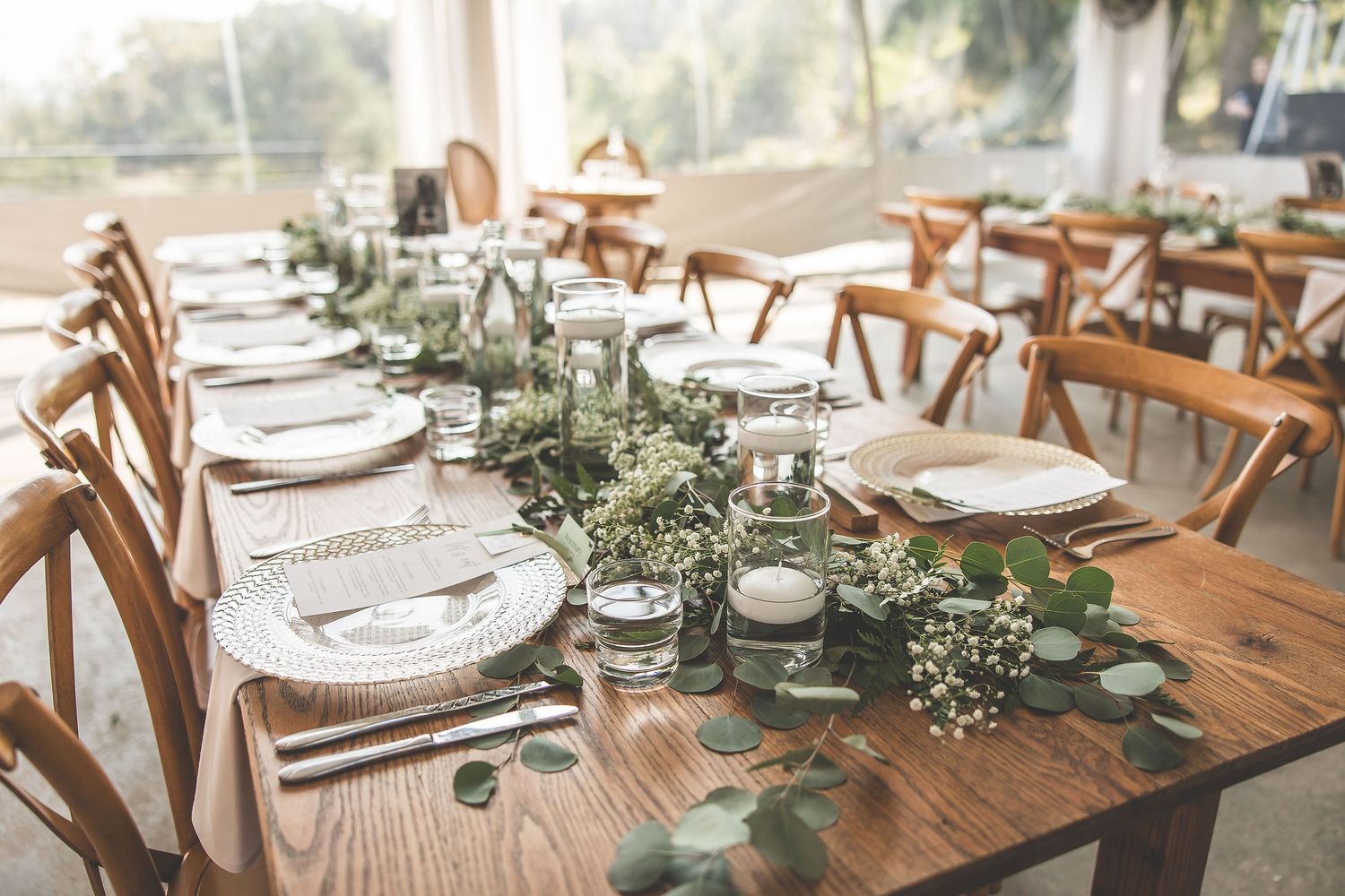 Rustic wedding reception tables set with greenery runners and wooden cross-back chairs.