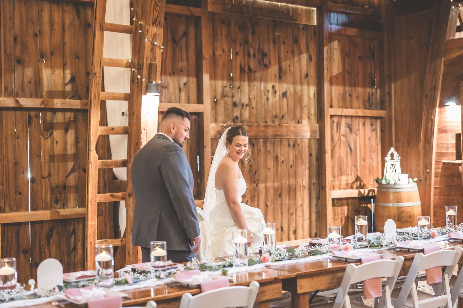 Wedding couple stands at rustic barn reception table with elegant place settings.