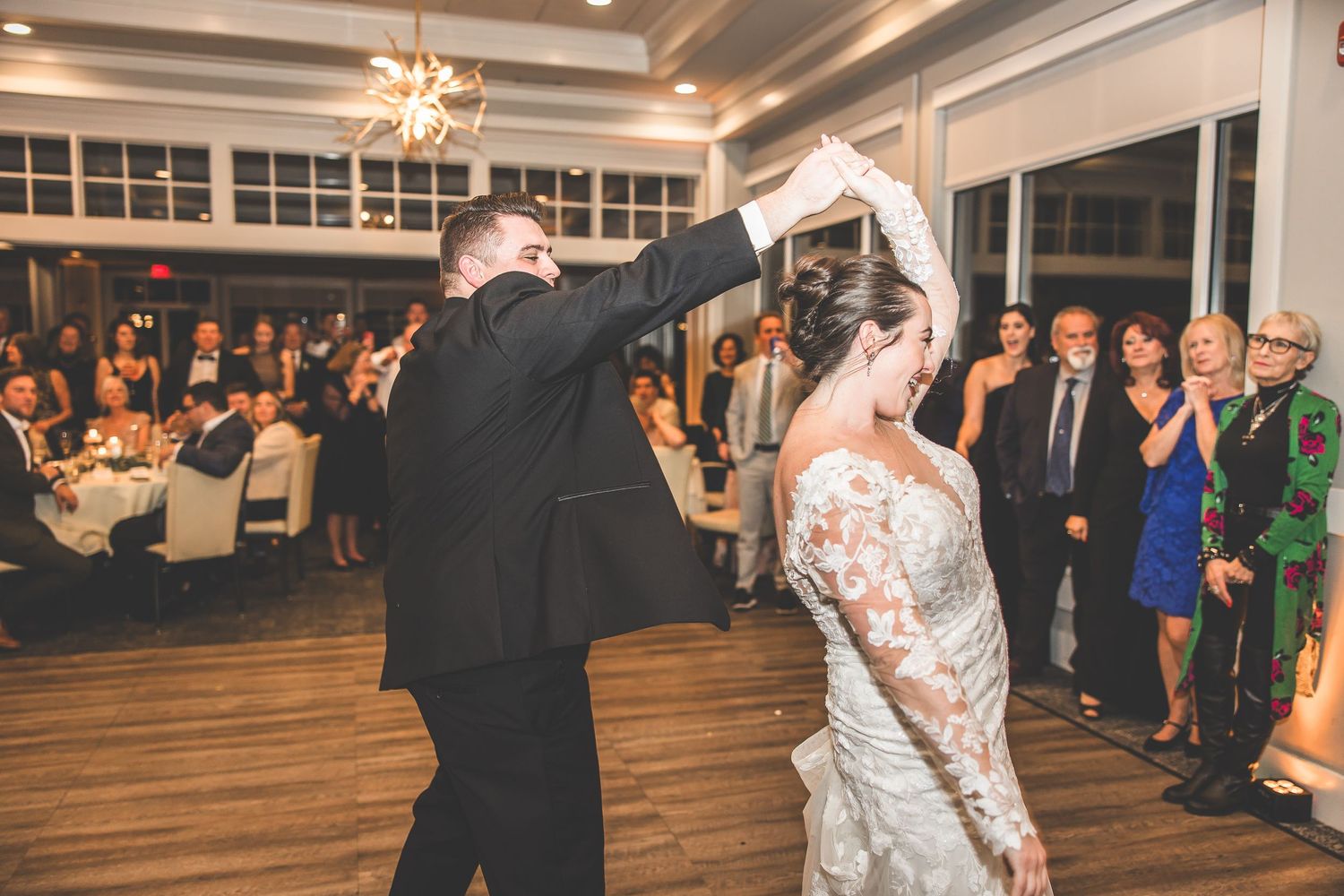 Wedding reception venue with wood floors and large windows filled with celebrating guests.