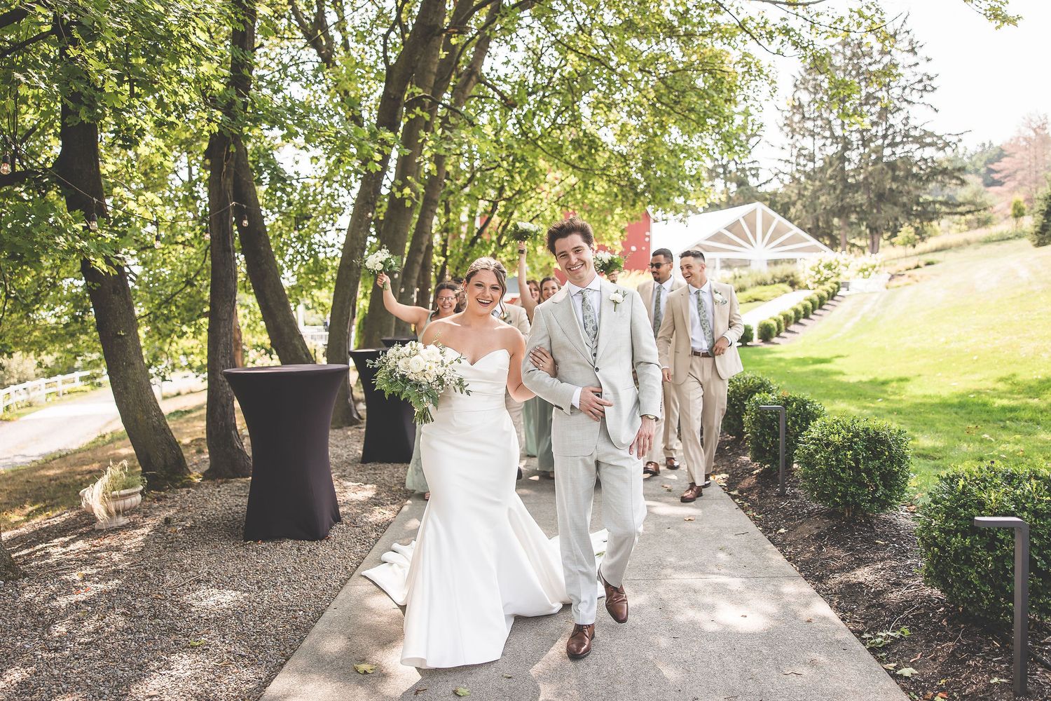 Wedding party walks together along a tree-lined path in coordinated sage and white attire.