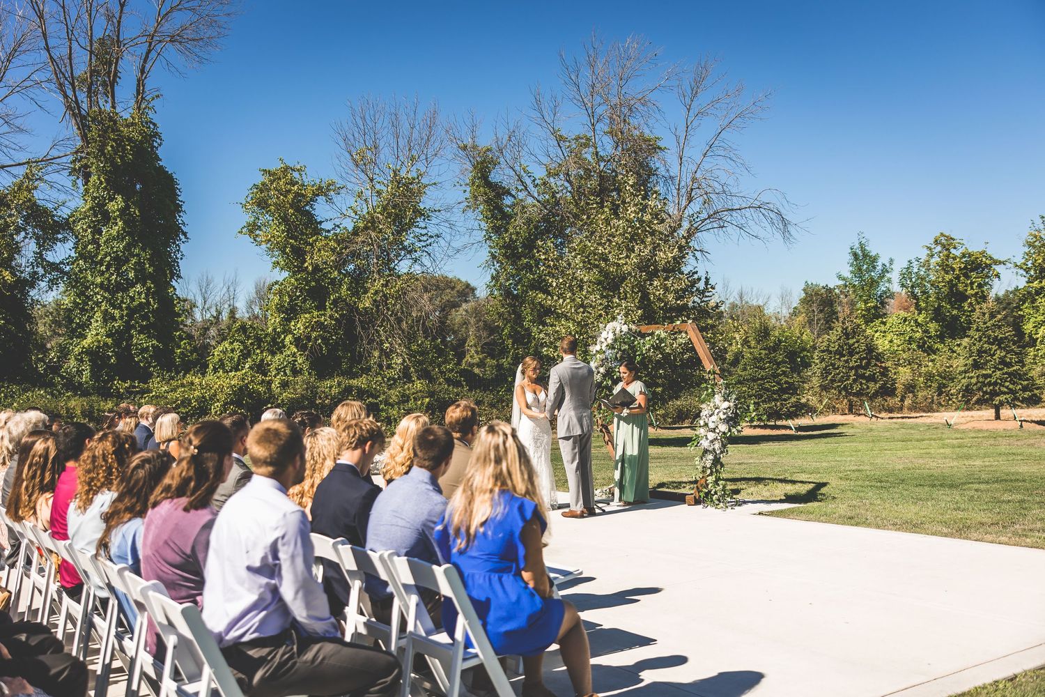 Outdoor wedding ceremony setup with white chairs and wooden arch decorated with flowers on a sunny day.