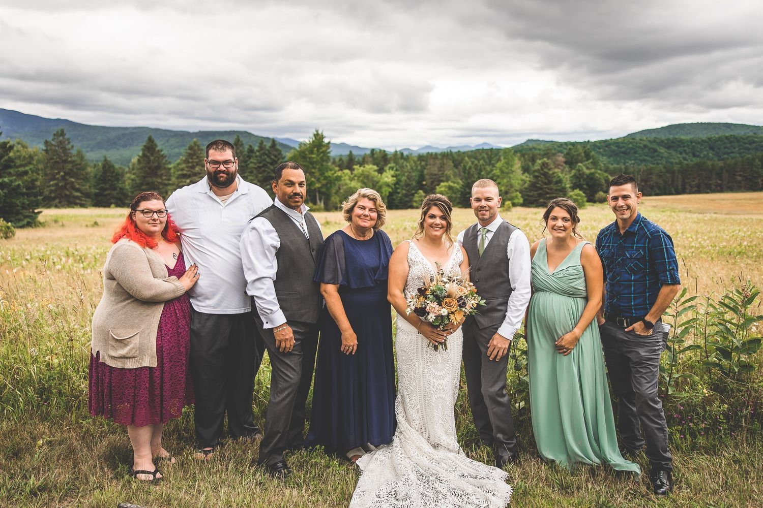 Wedding party poses together in a field with mountains and forest in the background.