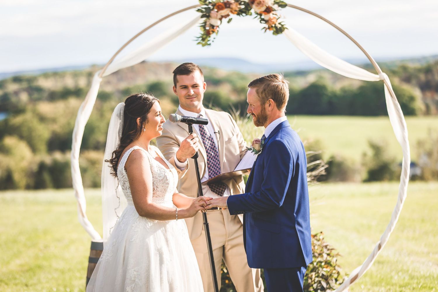 Floral arch decorated with peach roses stands against rolling hills during outdoor wedding ceremony.