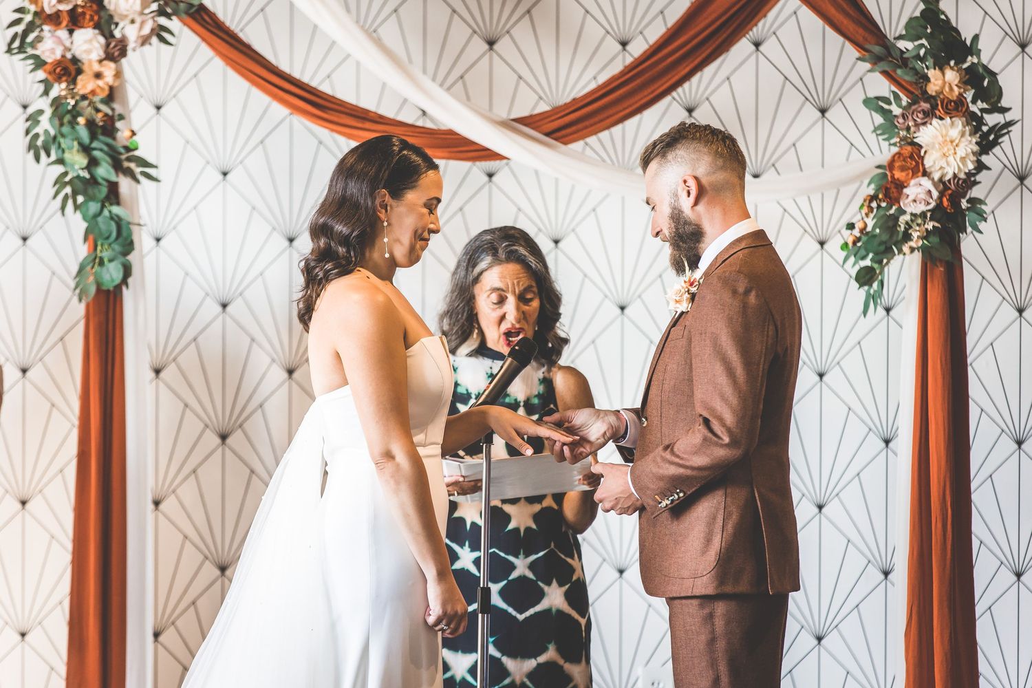 Wedding ceremony under wooden arch decorated with floral arrangements and copper draping.