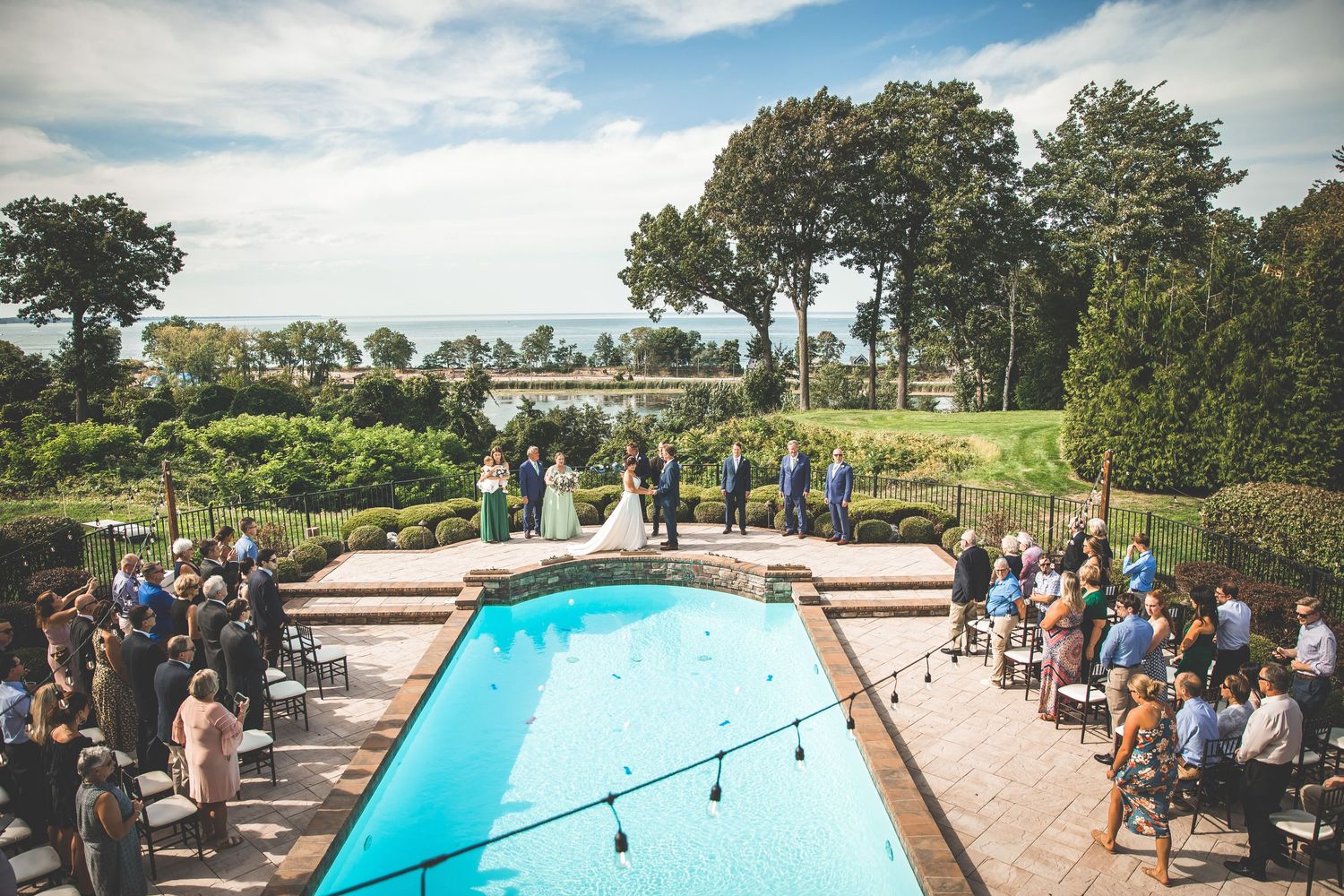 Panoramic view of wedding venue with swimming pool and guests gathered for outdoor ceremony.