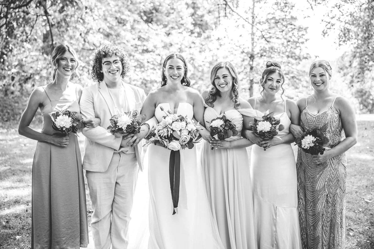 Wedding party in neutral tones poses together outdoors under trees in natural lighting.